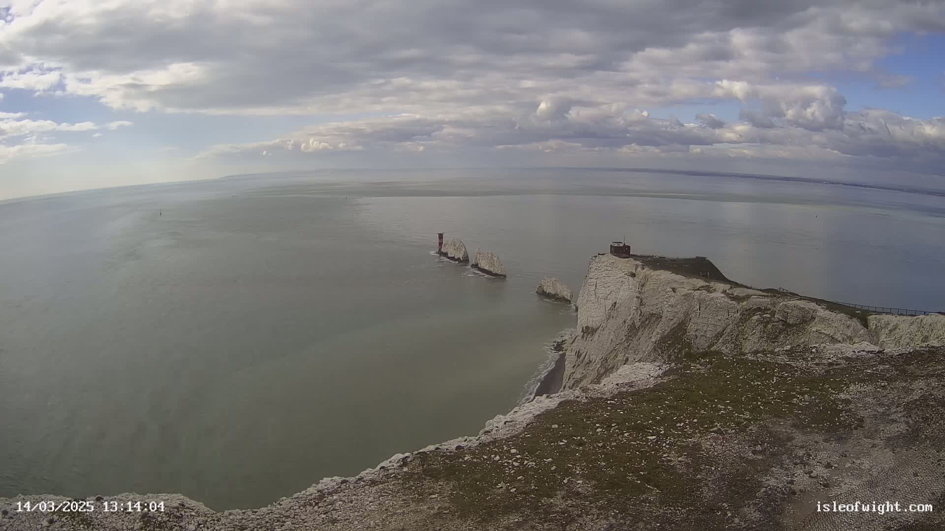 White cliffs overlook a calm sea with several stacks and a lighthouse under a partly cloudy sky.