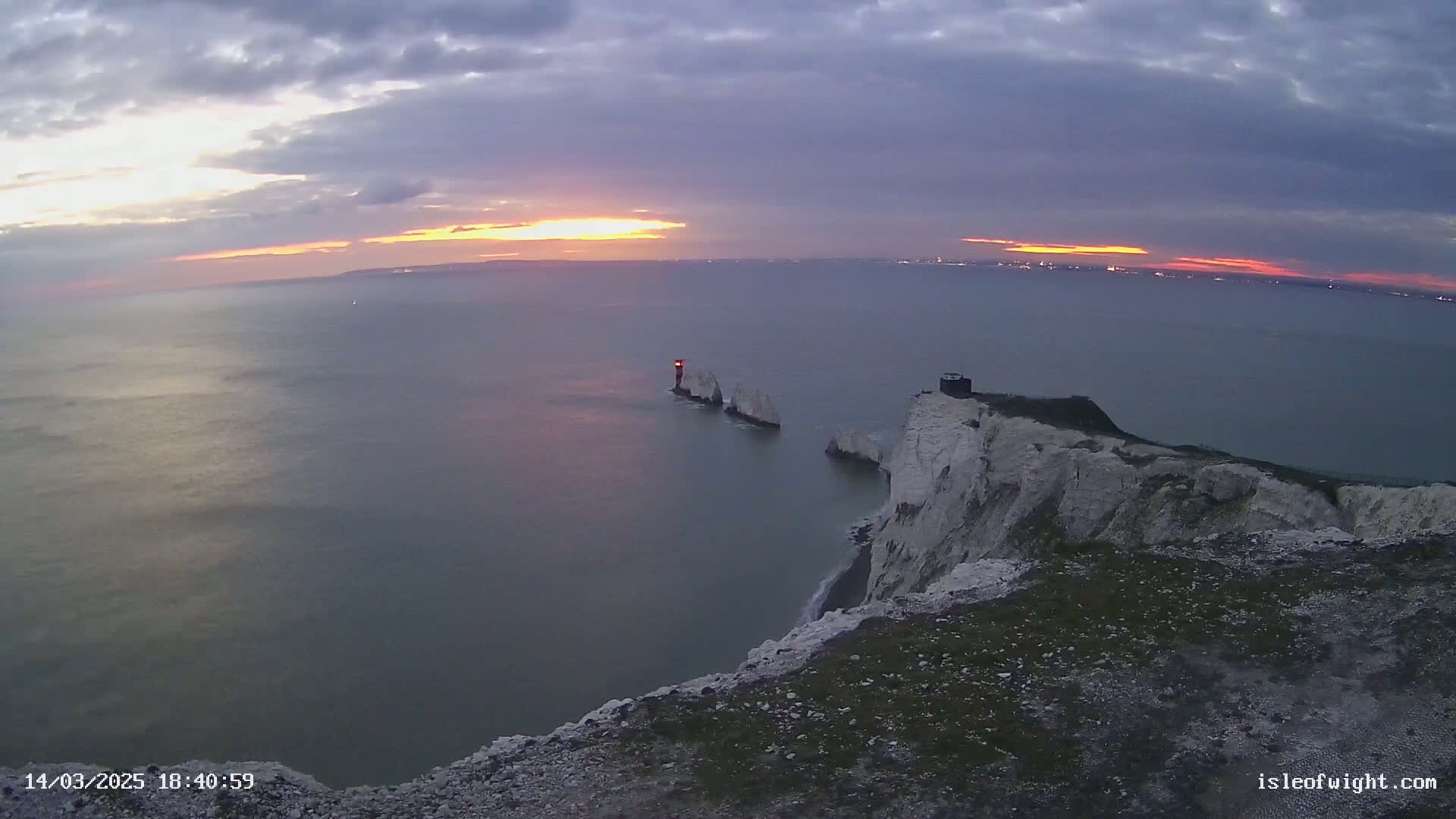 White cliffs overlook a calm sea at sunset, with a small island and lighthouse visible in the distance under a partly cloudy sky.