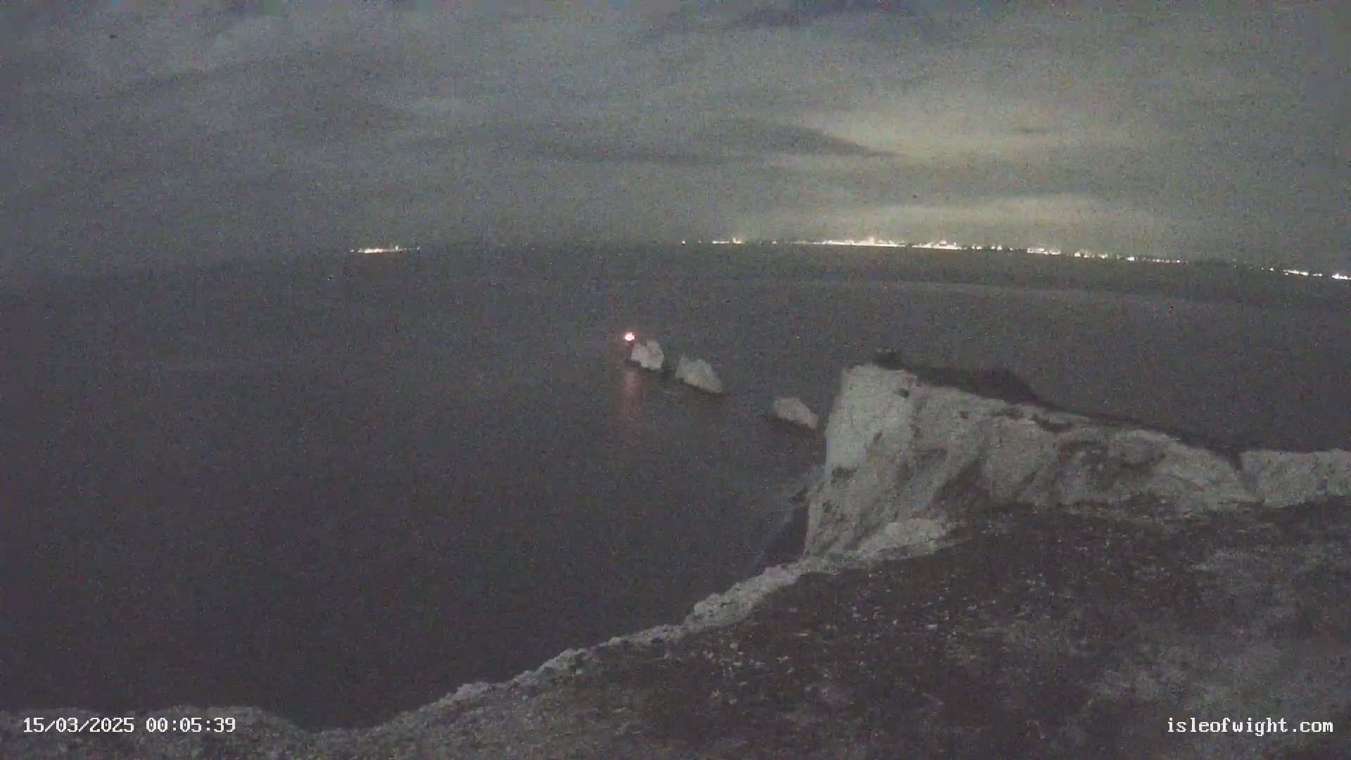 A nighttime view from a cliff overlooking a dark sea with white rocks and a faint light on the horizon.