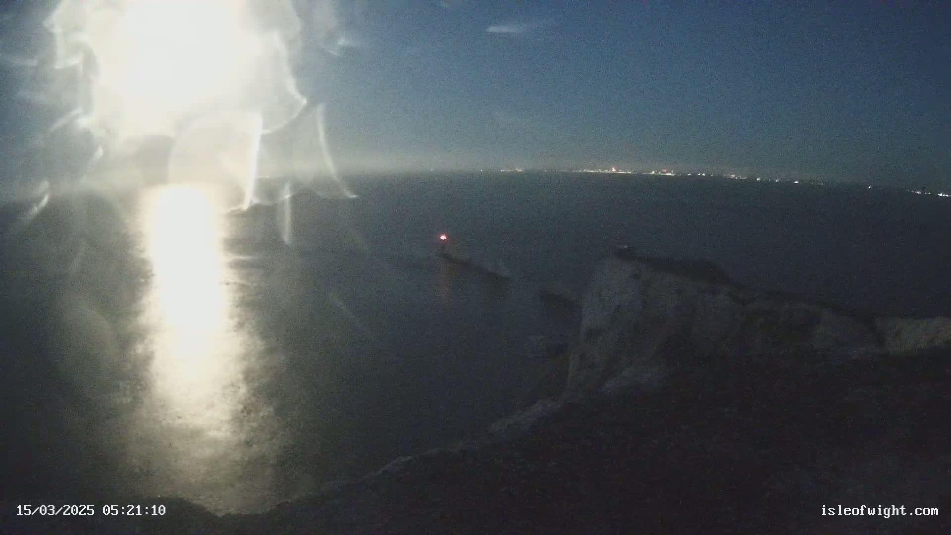 A nighttime coastal scene shows a bright light source in the foreground, a dark body of water with a distant light on the horizon, and a rocky cliff in the lower right.