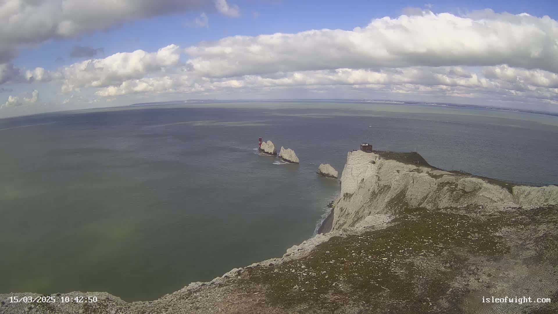 White cliffs overlook a calm sea with three stacks and a small building under partly cloudy skies.