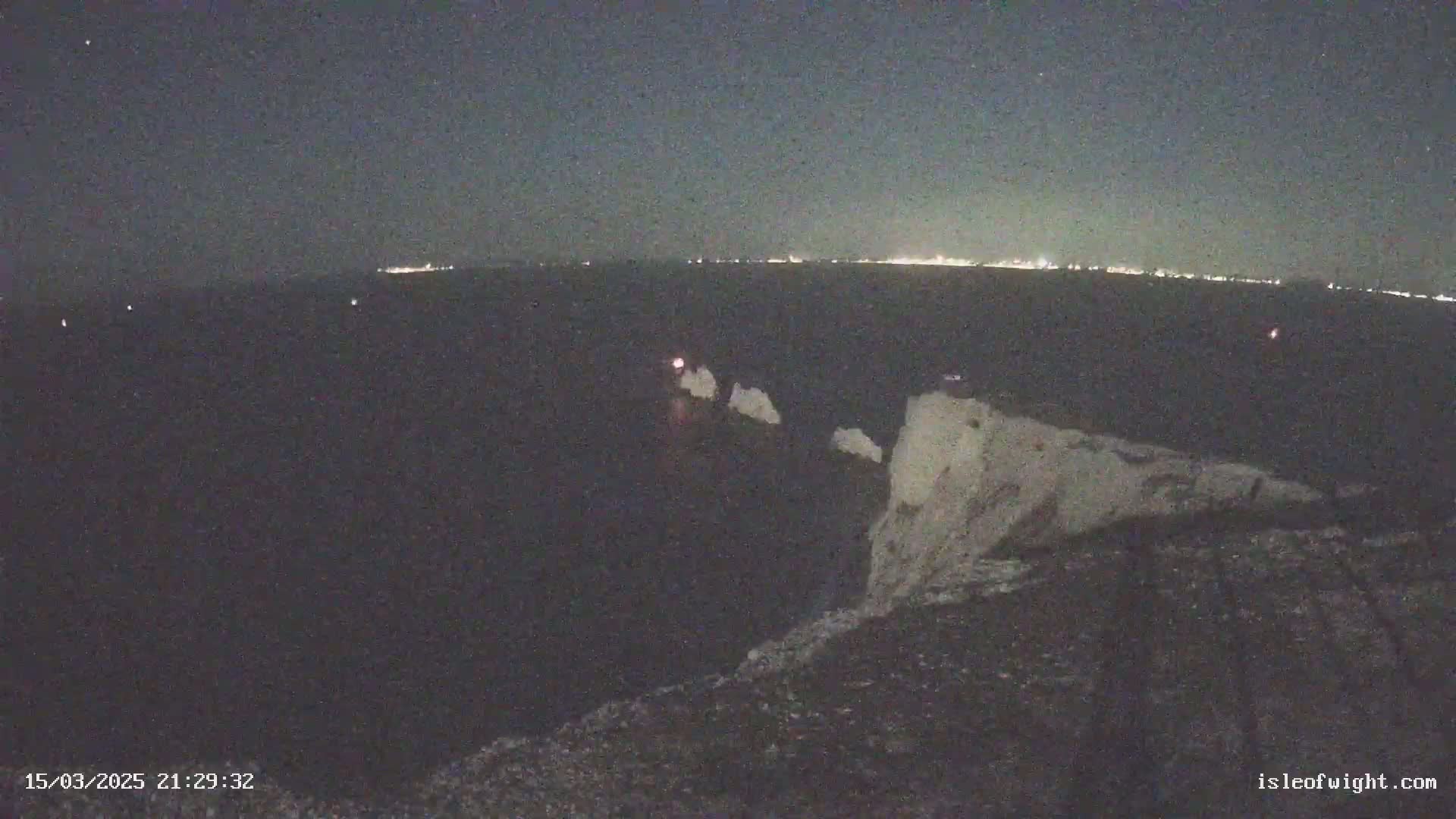 A nighttime, low-light view shows a cliff edge overlooking dark water, with a distant city skyline faintly illuminated on the horizon.
