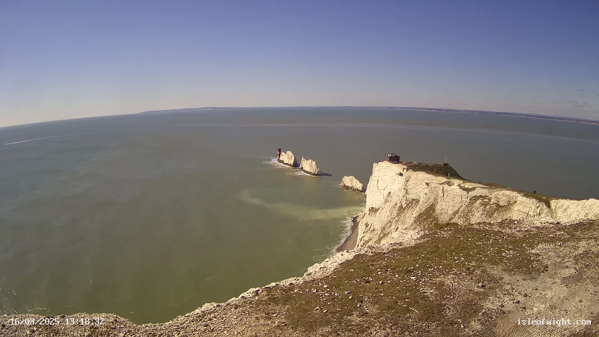 A high-angle view shows white cliffs overlooking a calm, greenish-grey sea with three white rock formations and a small red structure on one of them, under a clear blue sky.