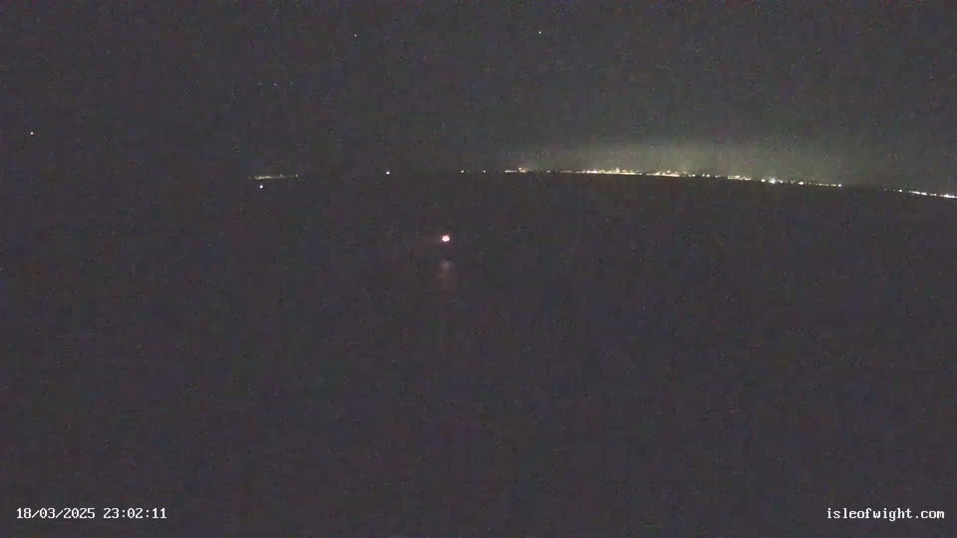 A dark, nighttime scene shows a body of water with a distant, faintly lit shoreline under a clear, starlit sky, and a single small light is visible on the water.