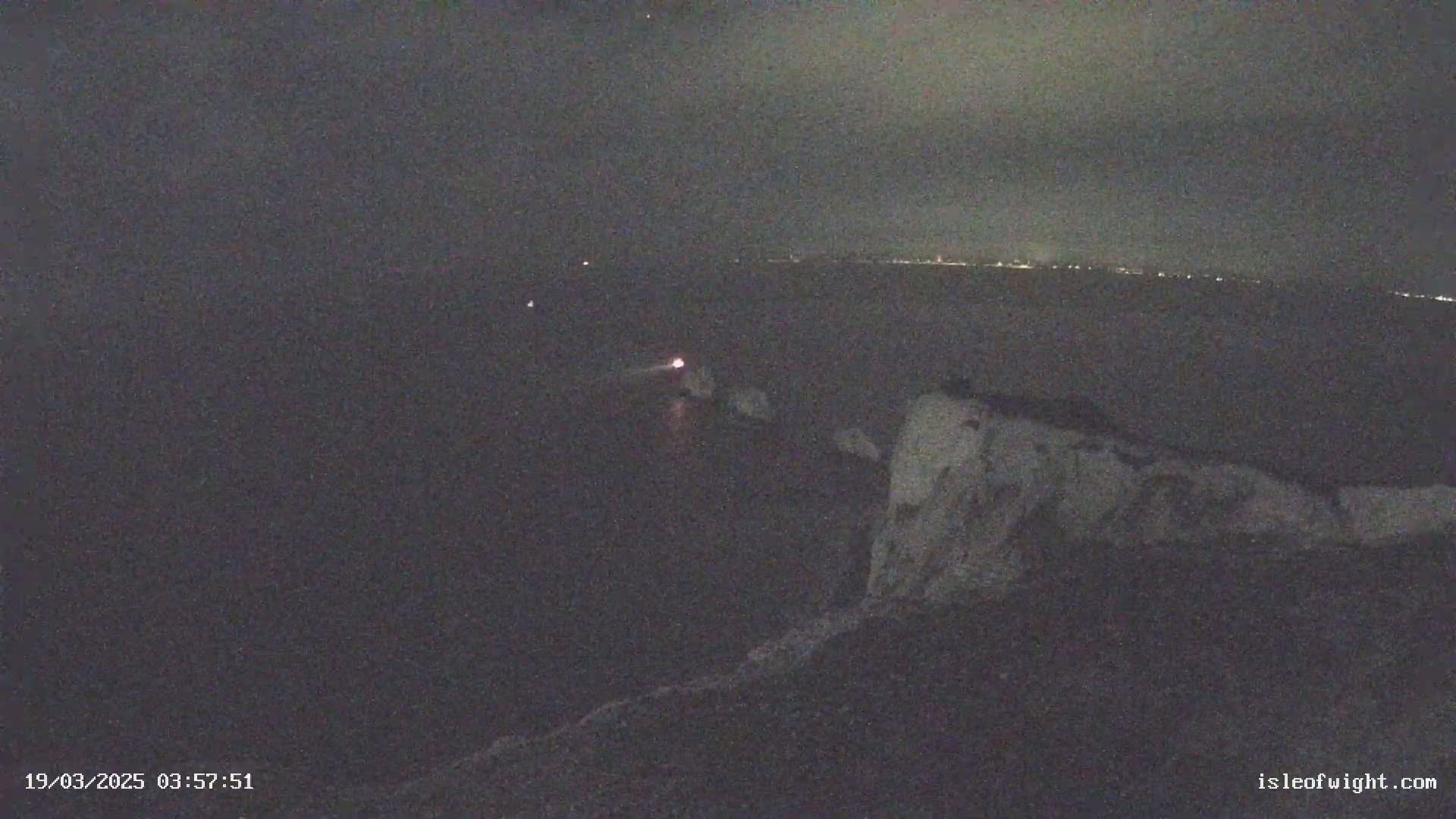 A nighttime view shows a dark body of water with a faint light on the horizon and a light-colored cliff in the foreground under a dark, cloudy sky.
