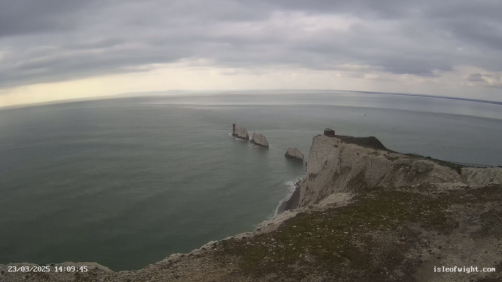 White cliffs overlook a calm, gray sea where several small, rocky islands are visible under a cloudy sky.