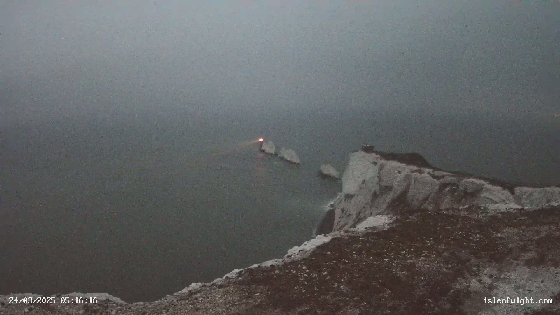 A hazy, dark grey day shows a cliff overlooking a body of water with white rock formations and a small light on one of them.