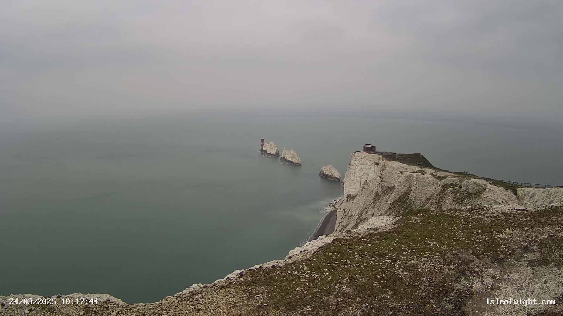 White cliffs overlook a hazy grey sea, with several smaller white rock formations and a small building visible in the distance.