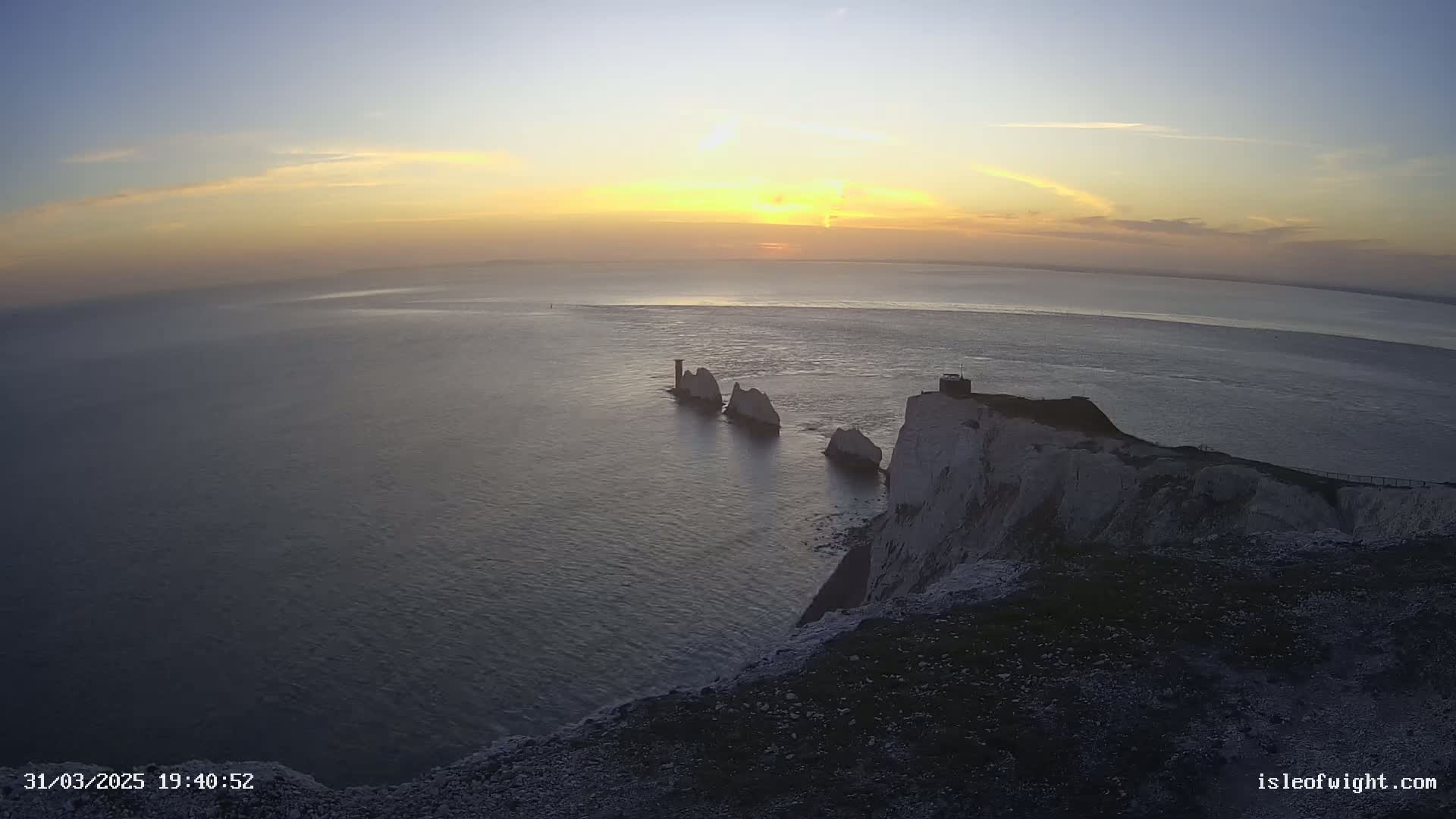 A sunset over a calm ocean, featuring white cliffs and several rock formations extending from the coast.