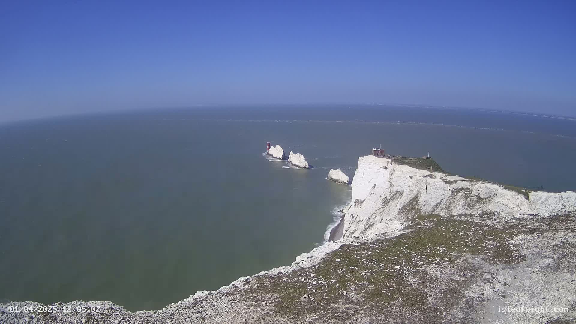White cliffs overlook a calm sea where three white rock formations with a red lighthouse are visible in the distance under a clear blue sky.