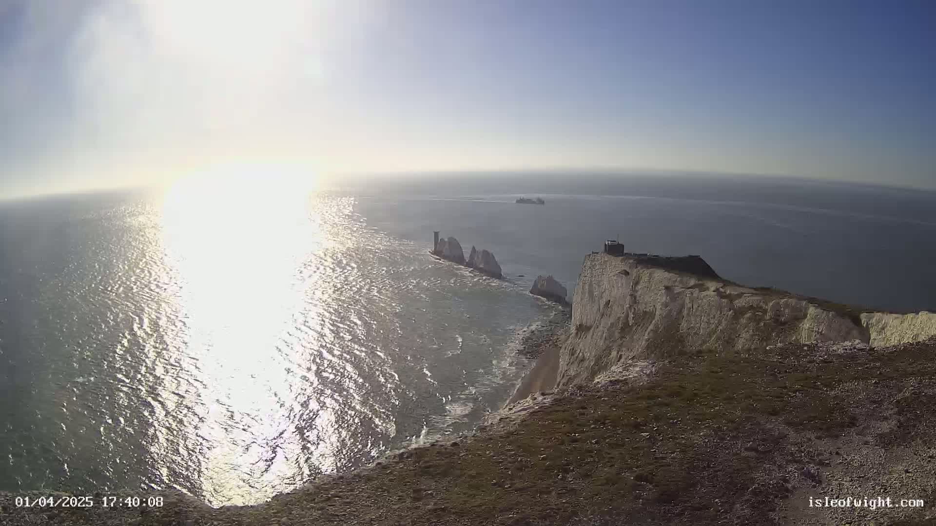 A sunny day reveals white cliffs overlooking a sparkling ocean with distant rocky formations and a ship on the horizon.