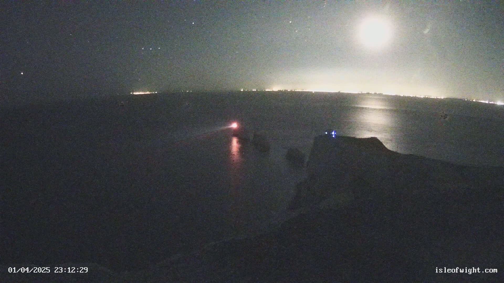 A nighttime coastal scene under a bright moon shows a dark body of water reflecting the moon's light, a distant shoreline with faint lights, and a dark cliff in the foreground with a small light source near the water.