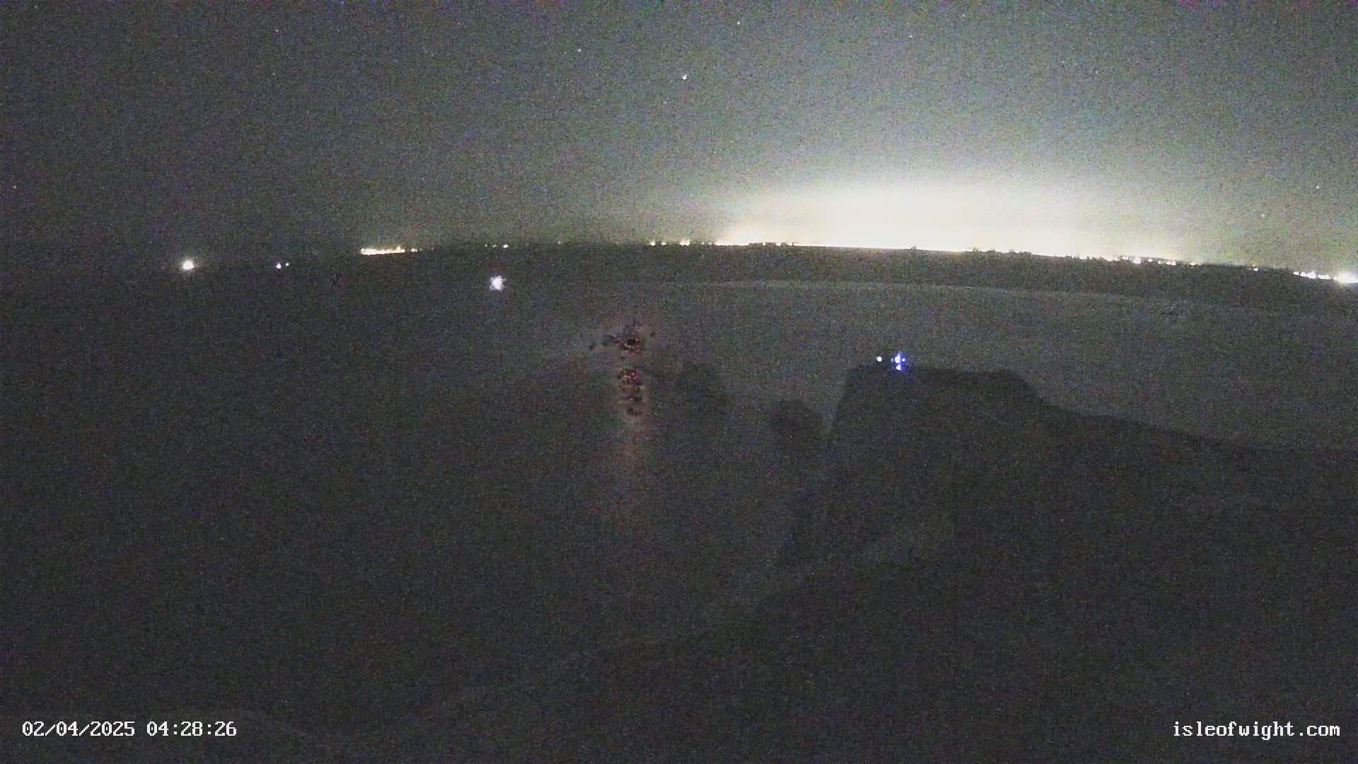 A nighttime long shot shows a dark landscape with a line of distant lights on the horizon and a cluster of faint lights in the middle ground, under a clear, starry sky.