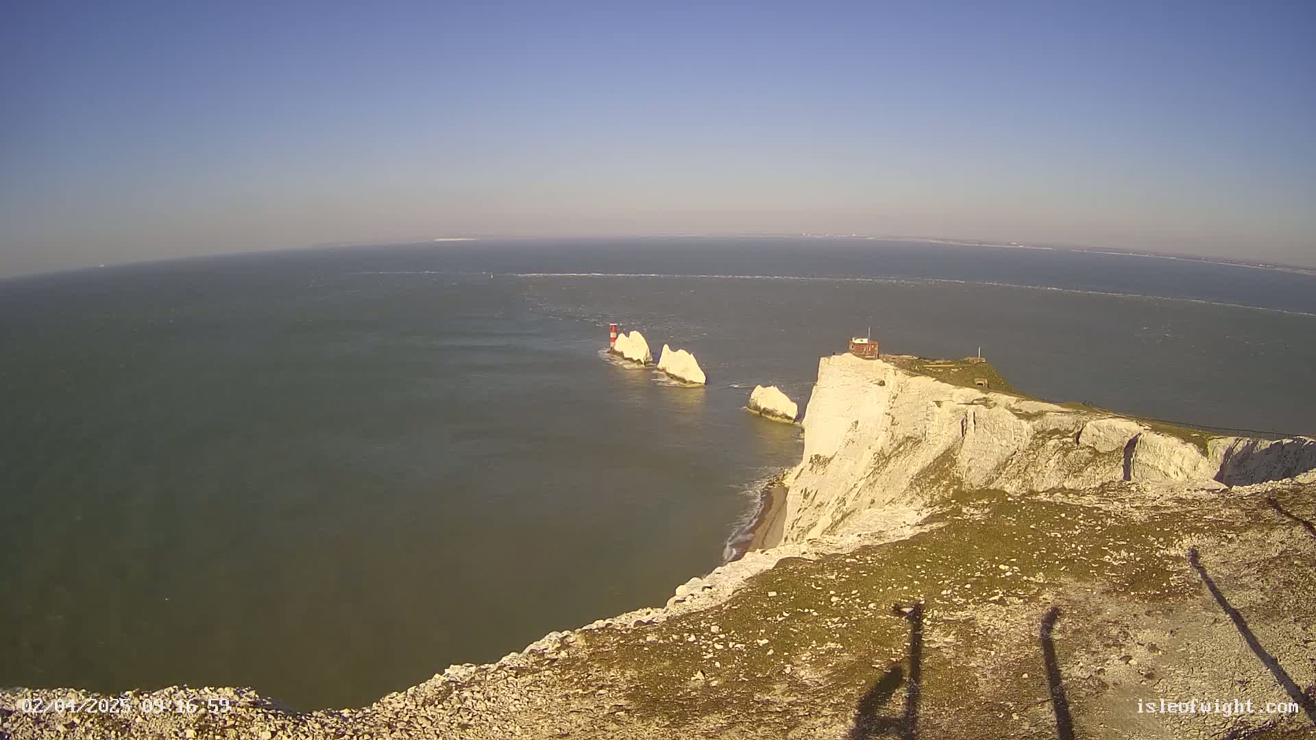 White cliffs overlook a calm sea with three white rock formations and a lighthouse in the distance under a clear blue sky.