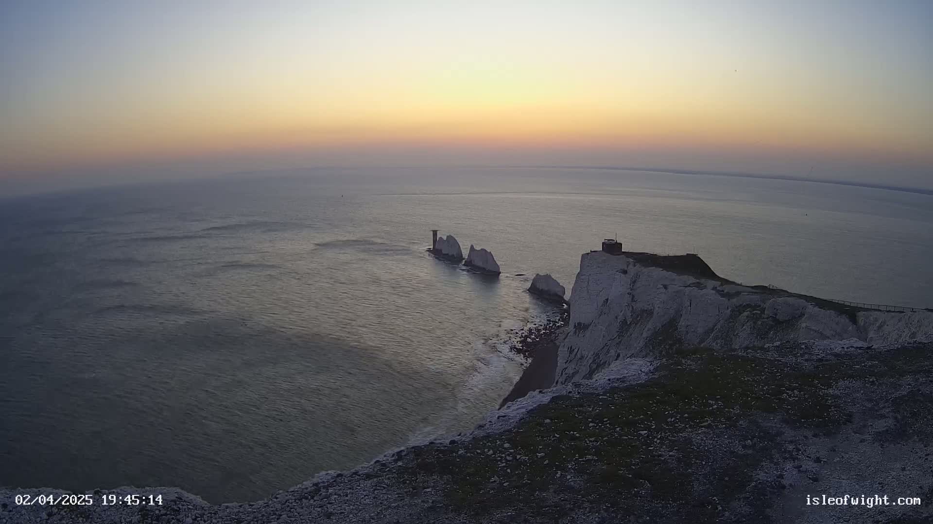 A high-angle view shows white cliffs meeting a calm sea at sunset, with several smaller white rock formations extending from the cliffs into the water.