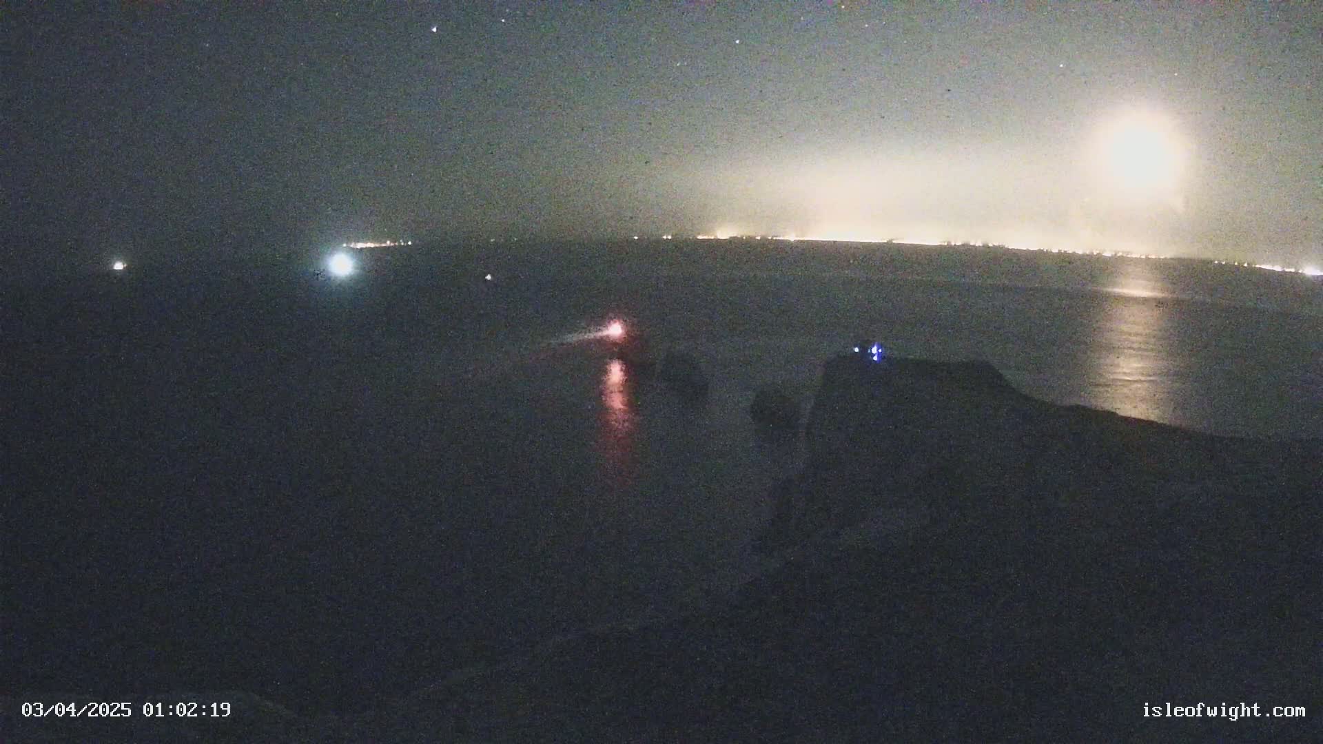 A nighttime view of a dark body of water with distant lights on the horizon under a partly cloudy sky.