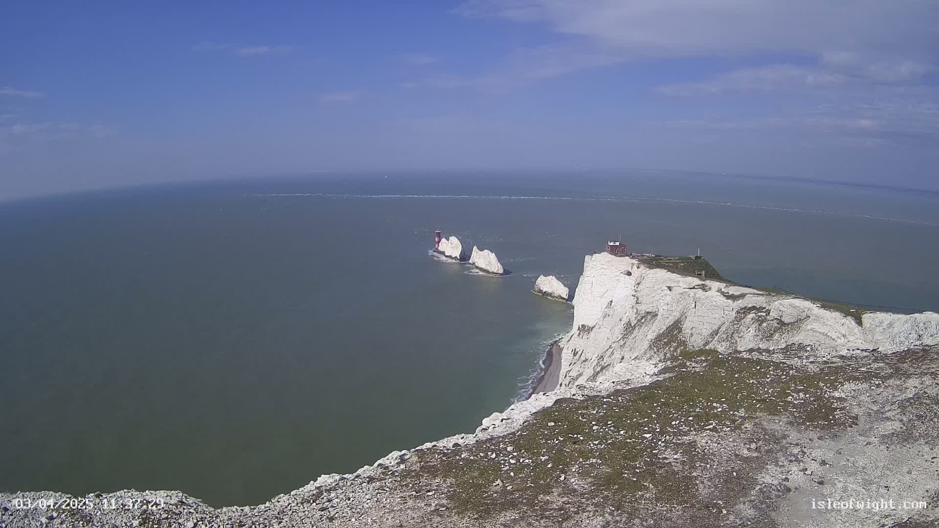 A high-angle view shows white cliffs meeting a calm sea under a mostly clear, bright blue sky; a line of smaller white rock formations and a lighthouse are visible in the sea.
