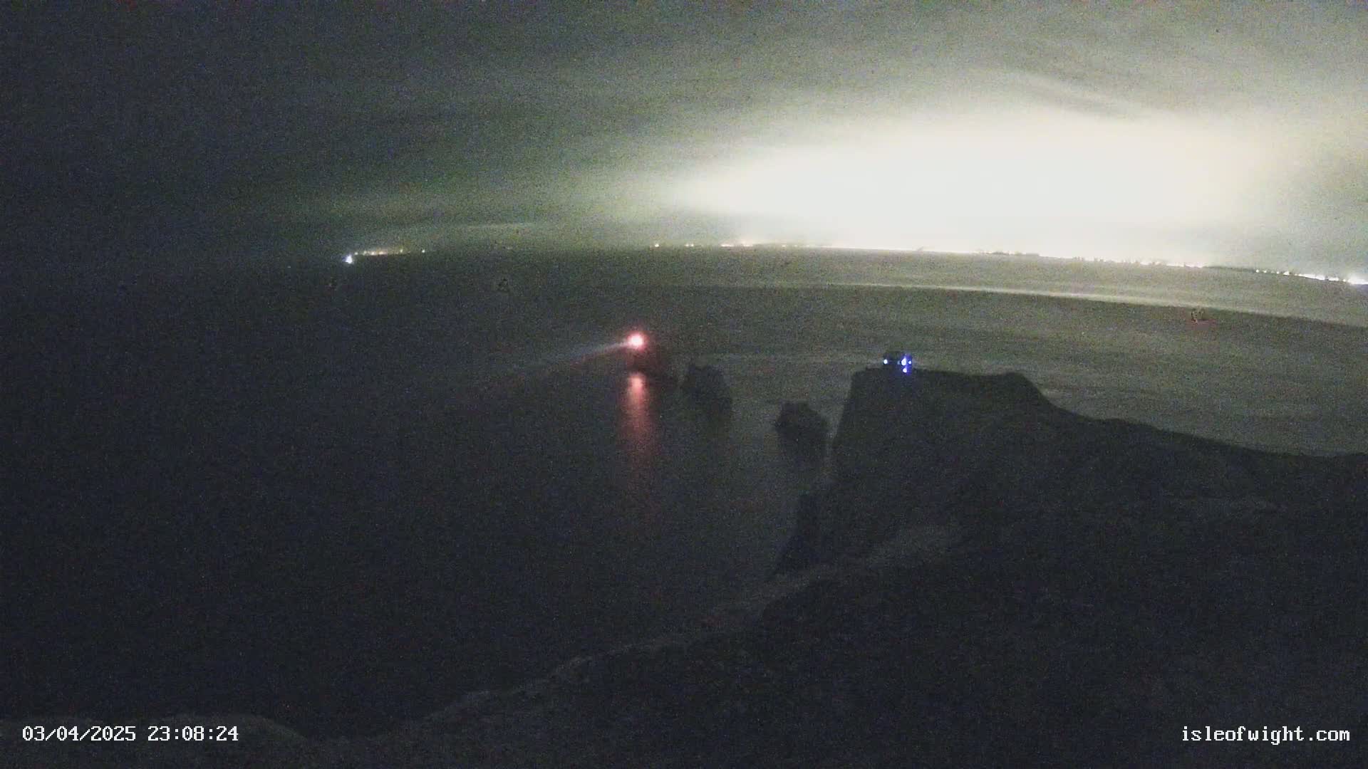A nighttime view of a dark body of water, with a distant, bright shoreline and a small light on a boat in the foreground.