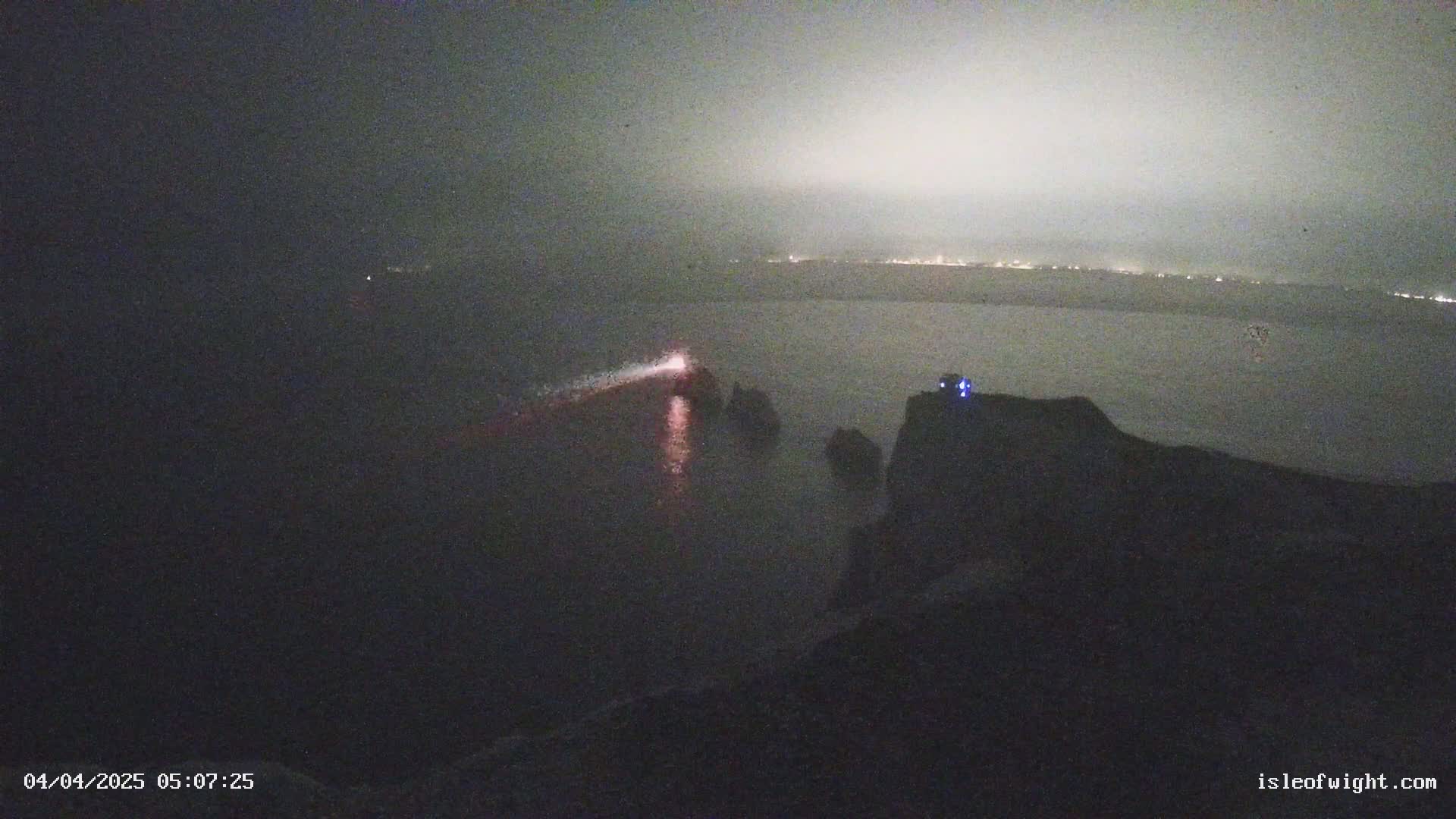 A nighttime coastal scene shows a body of water with a faint light source in the distance and several dark rocky outcrops in the foreground under hazy or foggy conditions.
