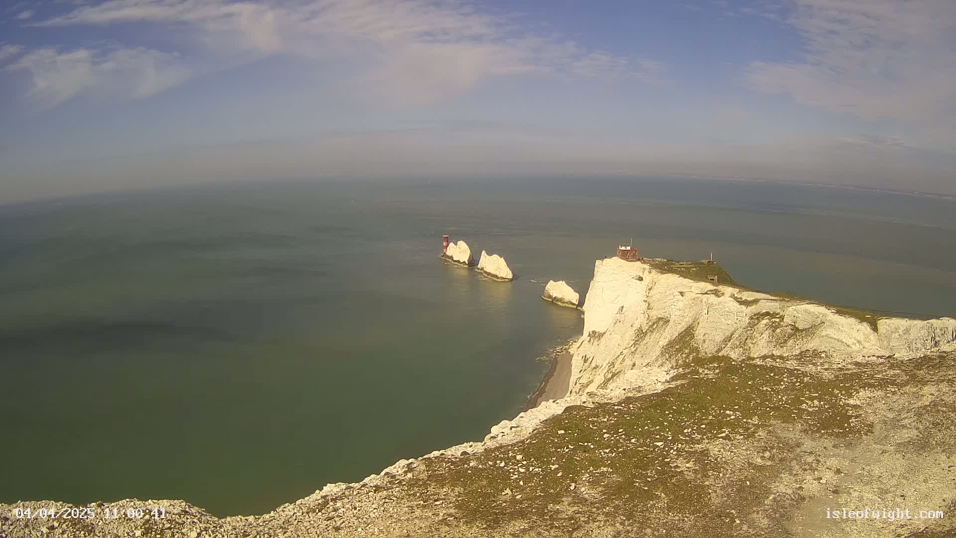 White cliffs overlooking a calm, green-blue sea with three white rock formations and a small building visible in the distance under a partly cloudy sky.