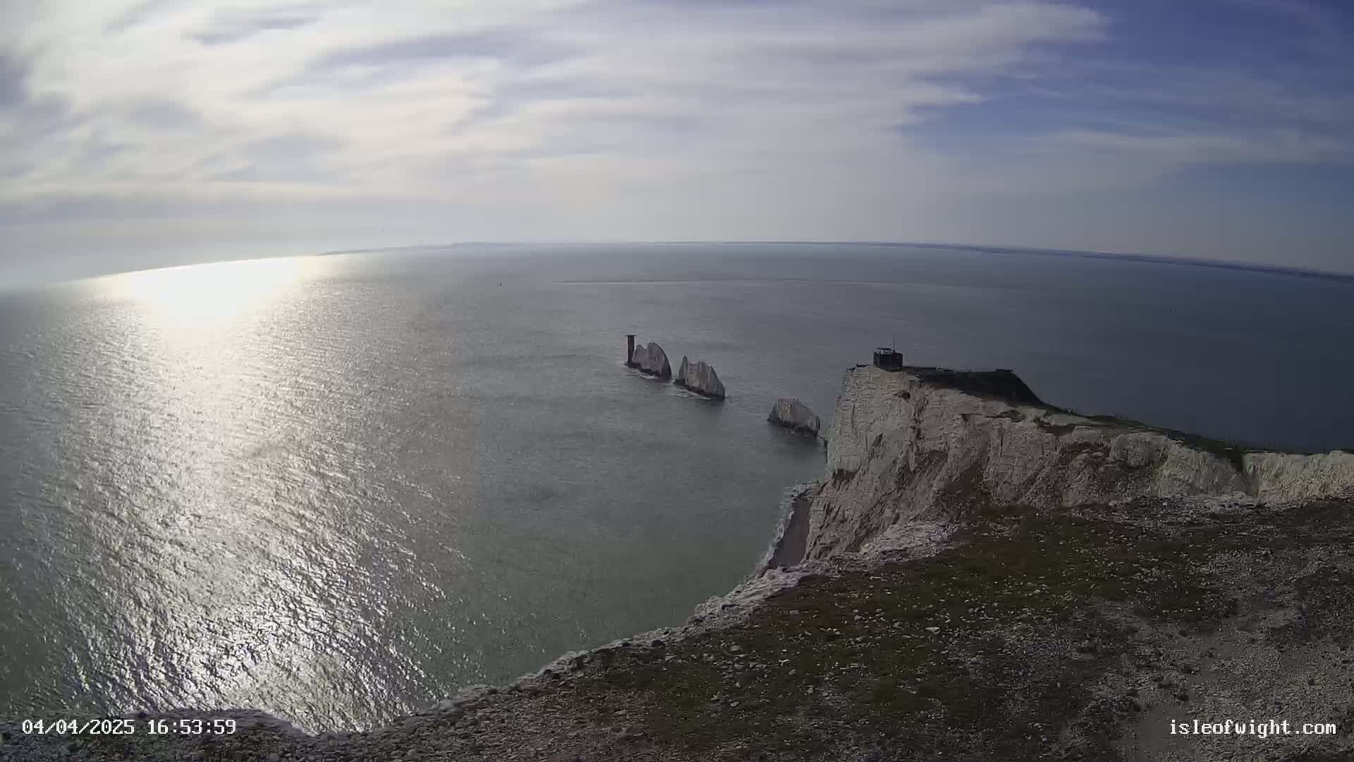 A high-angle, wide shot shows a bright, sunny day overlooking a calm sea with several white rock formations jutting out of the water near a tall white cliff.
