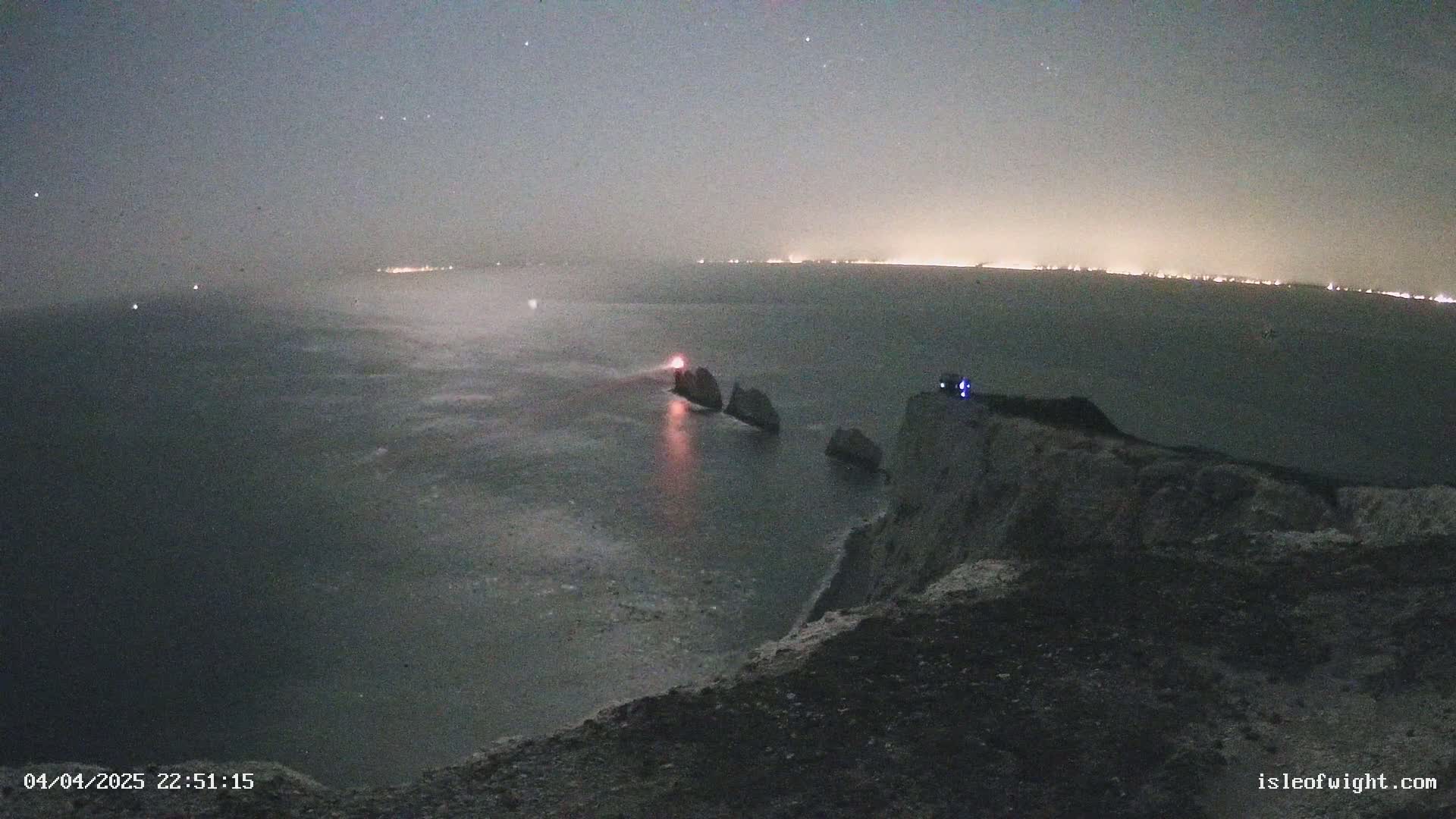 A nighttime, hazy view from a clifftop overlooking a dark sea with a lit-up lighthouse and distant lights on the horizon.