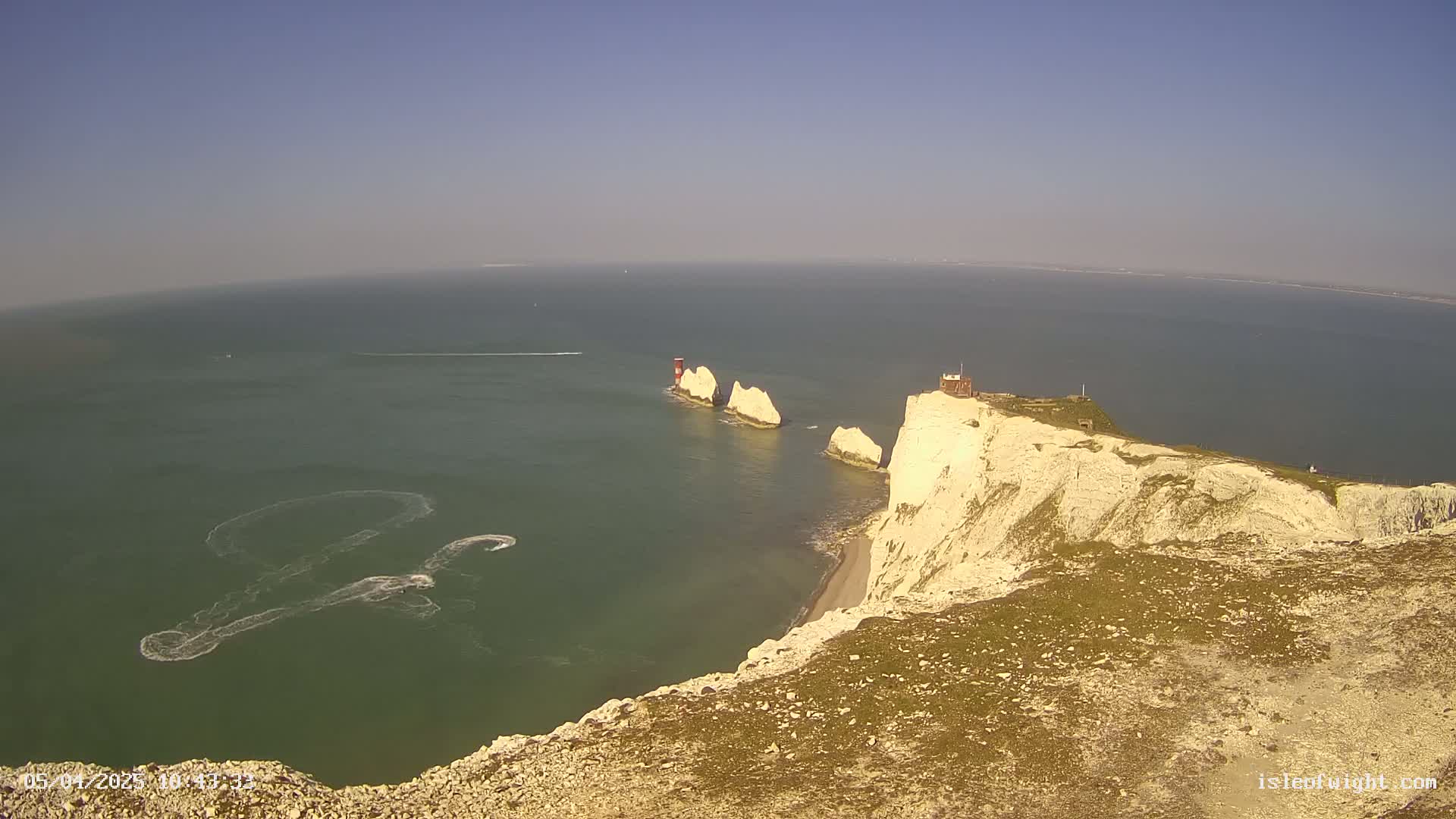 An aerial view shows white cliffs meeting a calm, green-blue sea where three stacks of white rock jut out from the water on a clear, sunny day.
