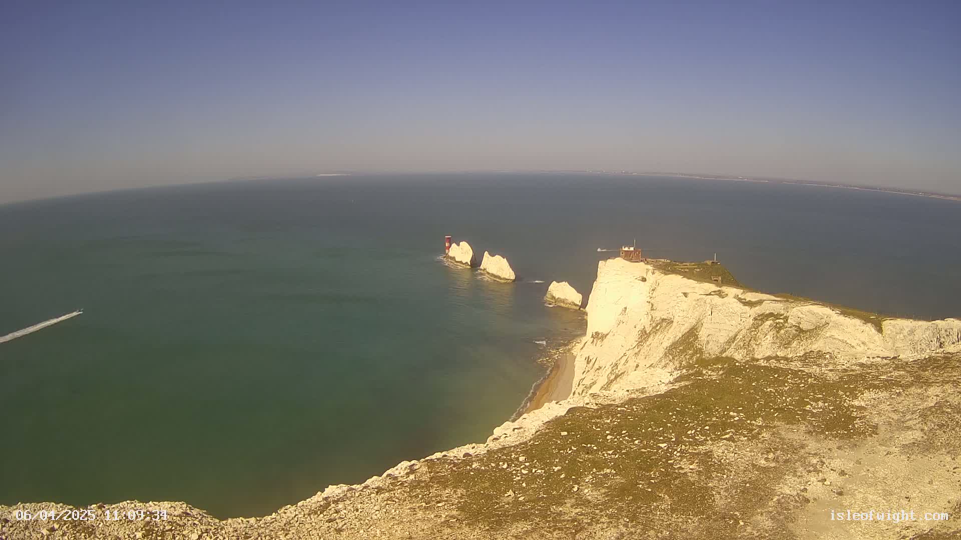 A high-angle view shows white cliffs meeting a teal sea, with two stacks of rock and a small building in the distance under a clear blue sky.