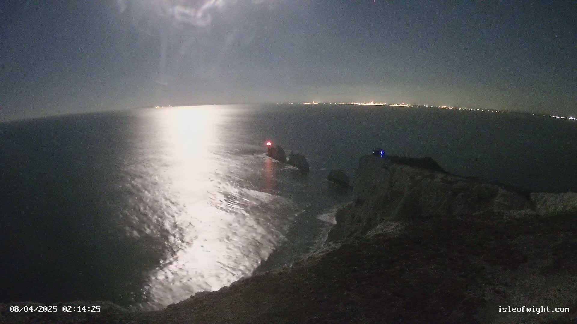 A moonlit night scene shows a rocky coastline with a body of water reflecting the moon's light, distant city lights visible on the horizon.