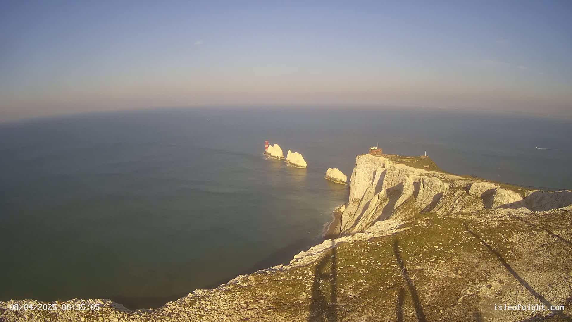 White cliffs overlook a calm sea with several smaller white rock formations and a small building visible in the distance under a clear sky.