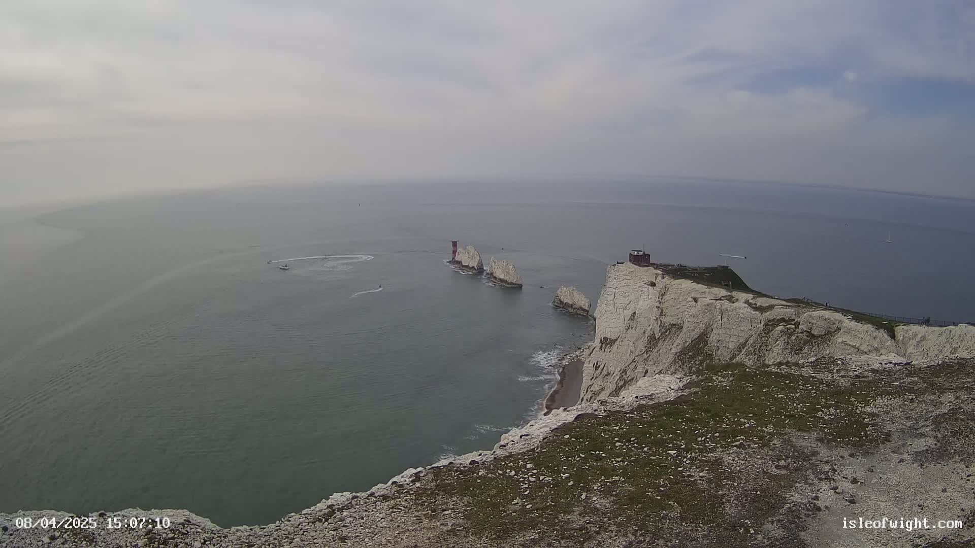 A high-angle view shows white cliffs overlooking a calm sea with a few small boats and a group of offshore stacks under an overcast sky.