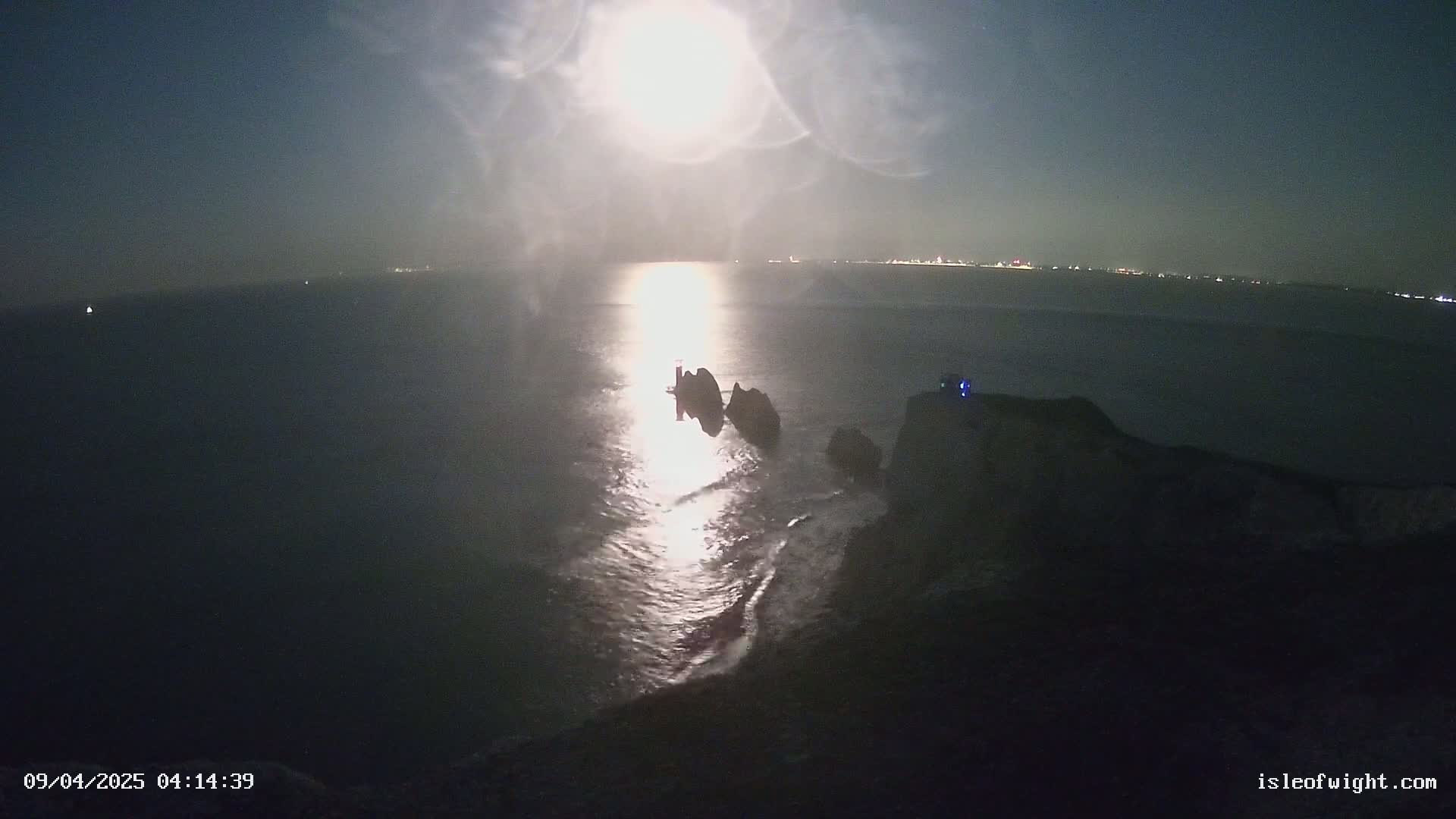 Two dark, rocky islands are partially visible in a moonlit sea at night, with a faint glow of distant lights on the horizon.