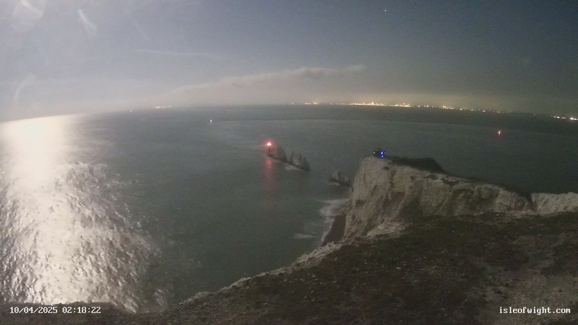A nighttime view from a clifftop shows a dark sea with a small group of rocks and a light visible offshore, under a mostly cloudy sky.