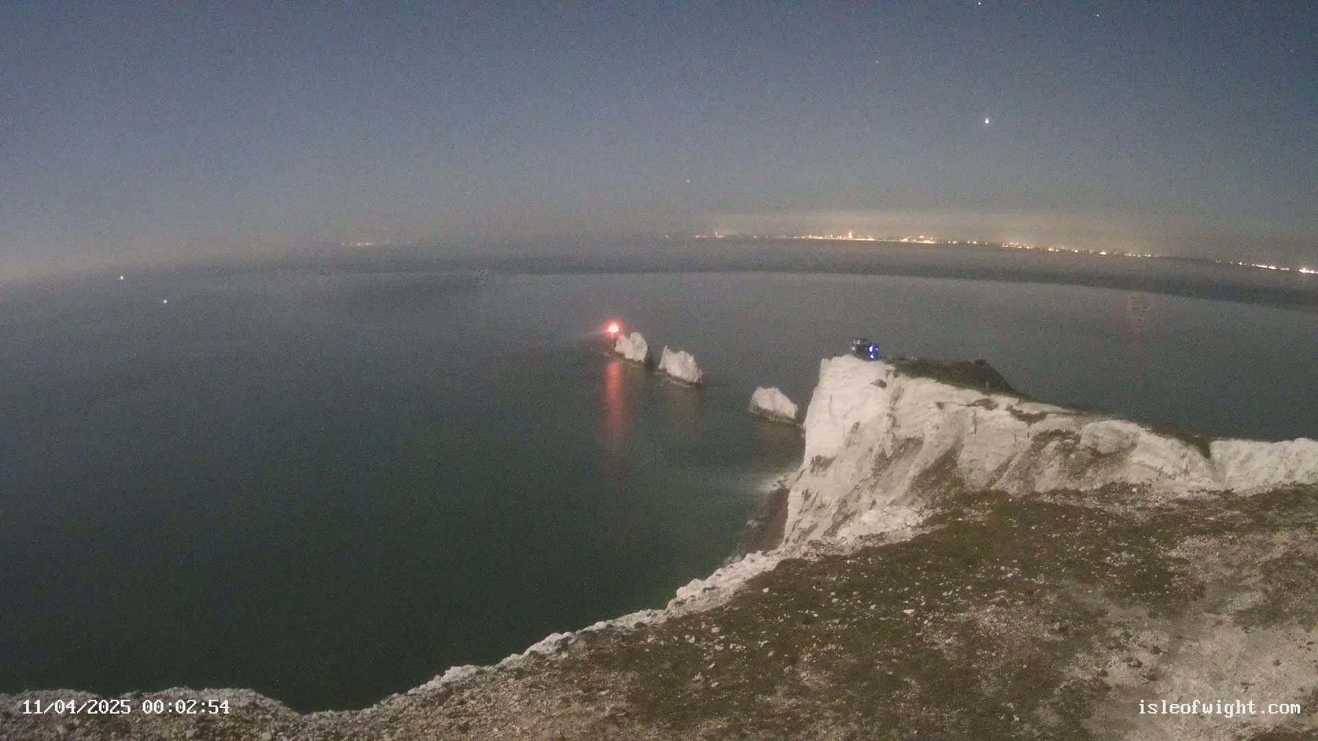 A nighttime coastal scene shows white cliffs overlooking a dark sea with three rock formations offshore, one illuminated with a red light, under a mostly clear sky with a few stars.