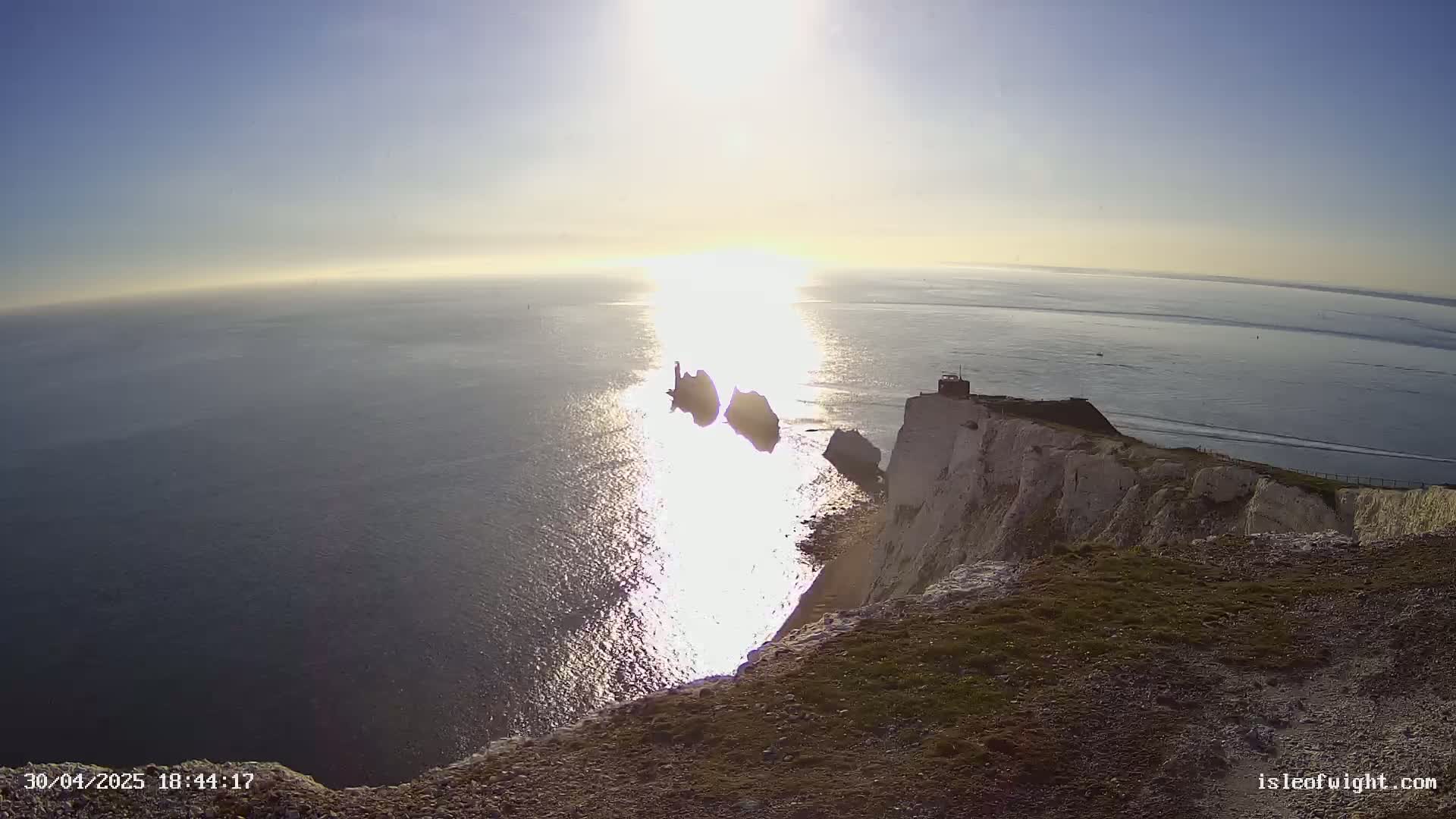 A bright, sunny day reveals a view from a clifftop overlooking the ocean, where two distant rock formations are visible in the glistening water.