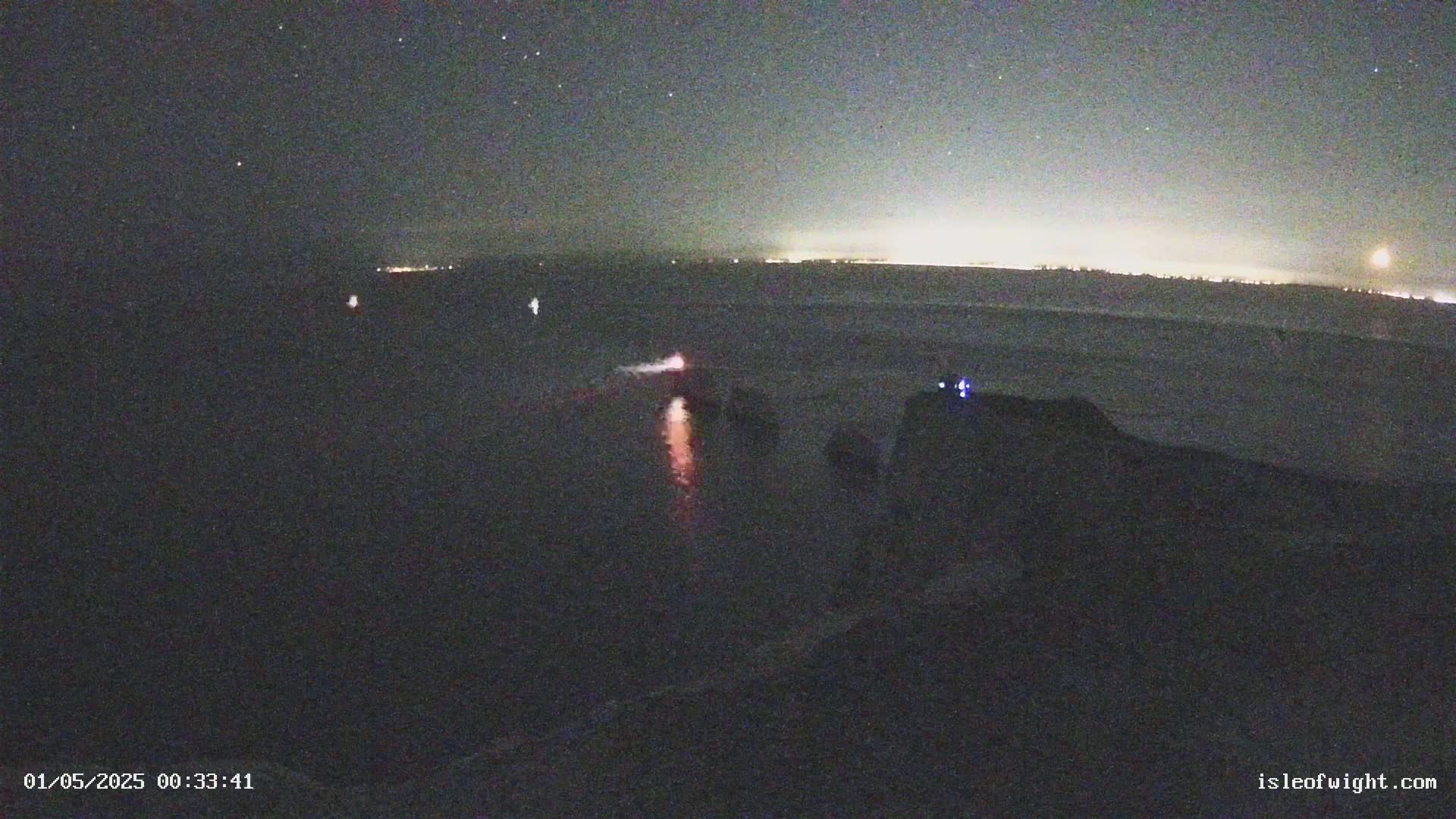 A nighttime view of a dark body of water with faint lights on the horizon and a reddish glow near the bottom.