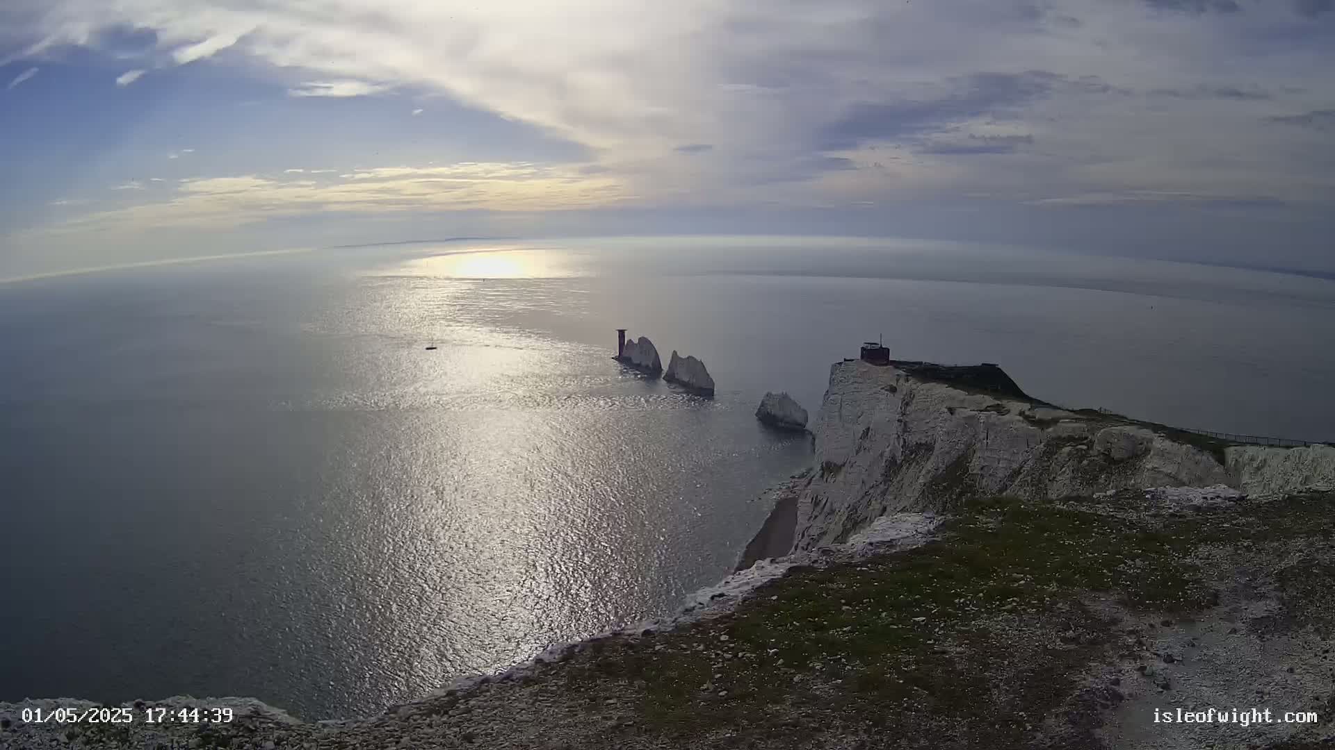 White cliffs overlook a calm sea reflecting sunlight, with several smaller rock formations visible in the distance under a partly cloudy sky.