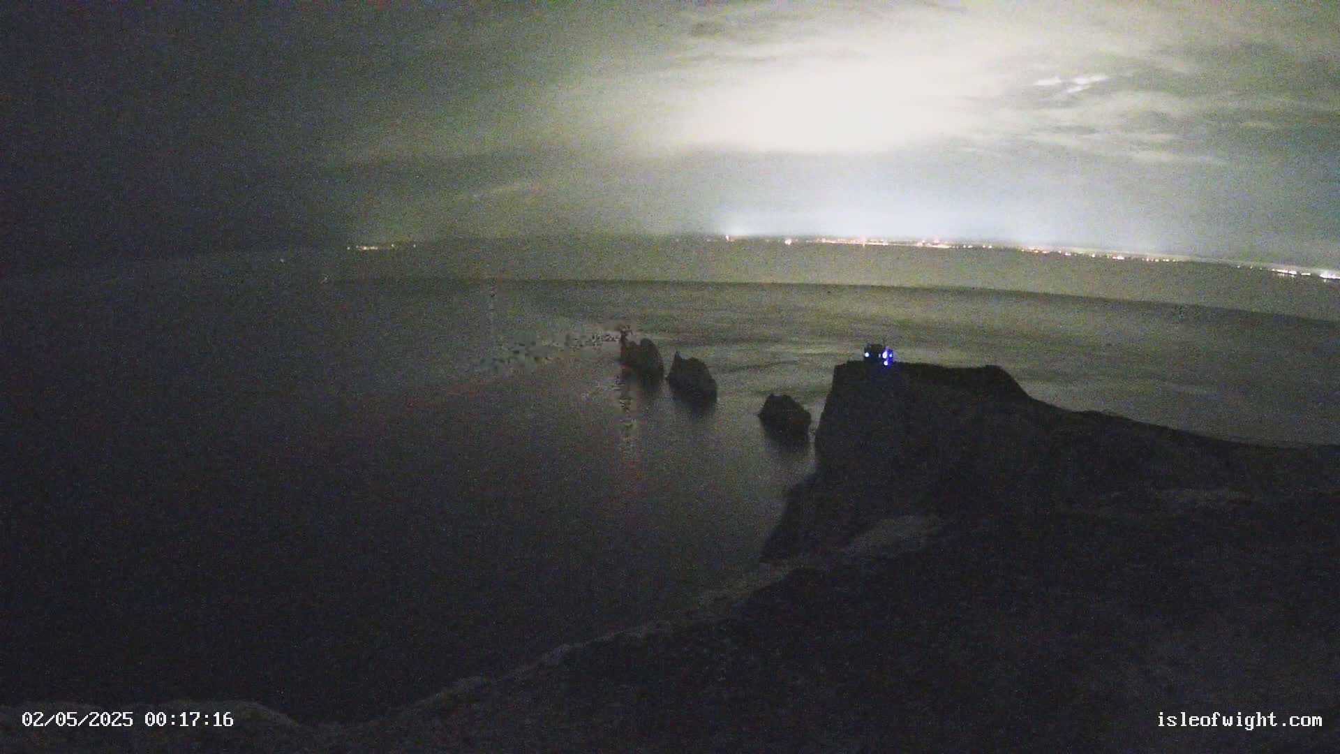 A nighttime coastal scene shows dark rocks and a body of water under a cloudy sky, with a distant shoreline faintly illuminated.