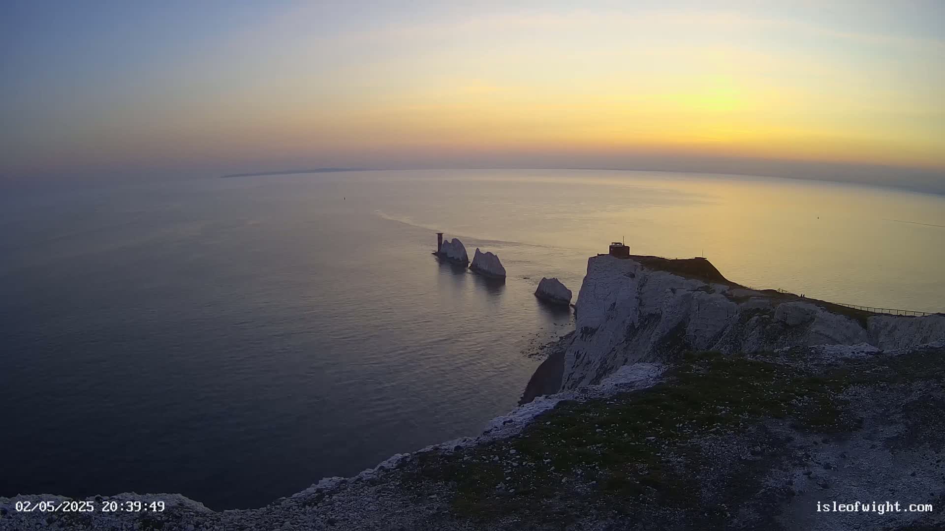 A high-angle view shows a rocky white cliff overlooking a calm ocean at sunset, with several small white islands visible in the water.