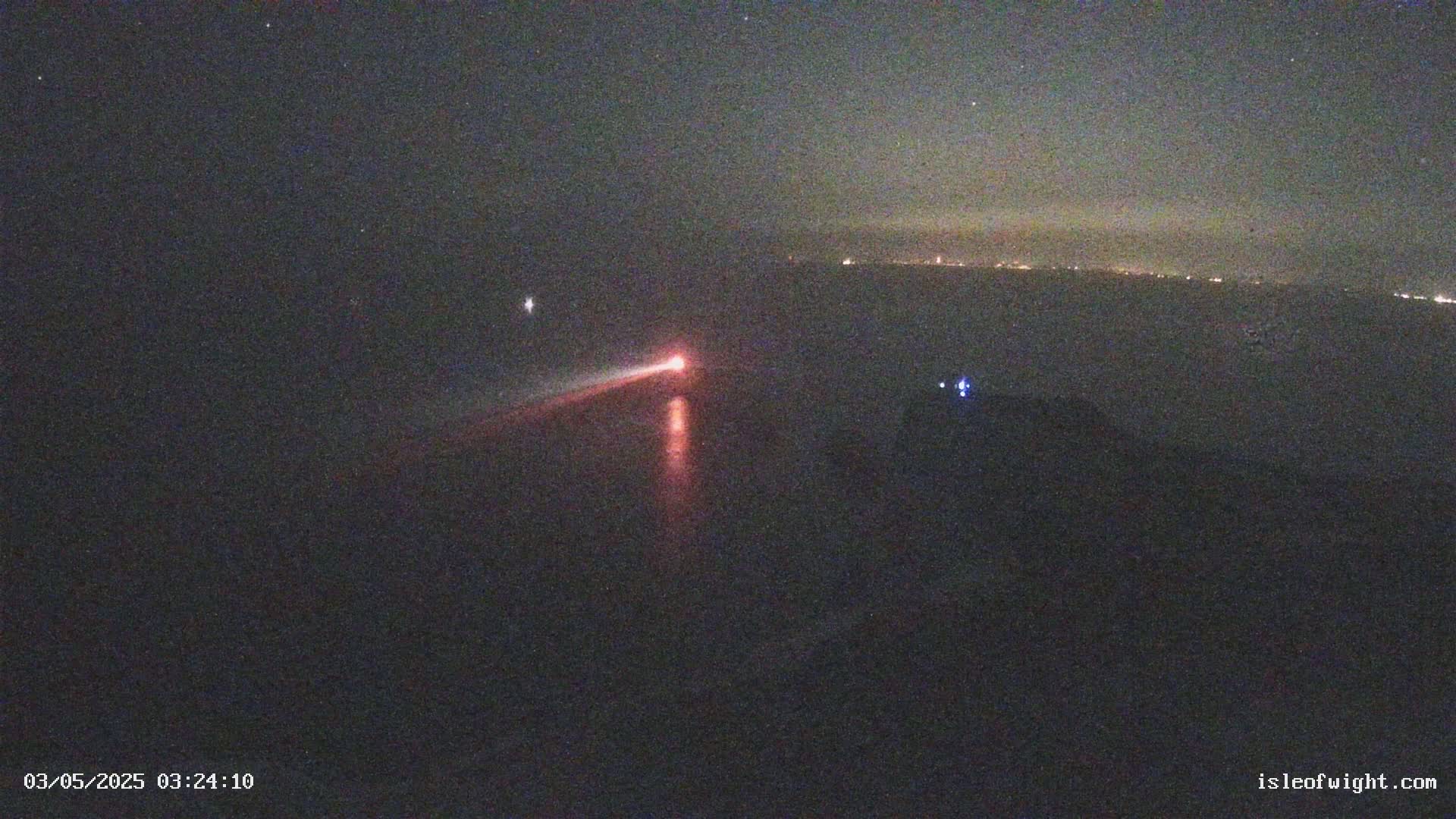 A nighttime long-exposure shot shows a red light streaking across a dark body of water, with a faint line of lights visible on the horizon.