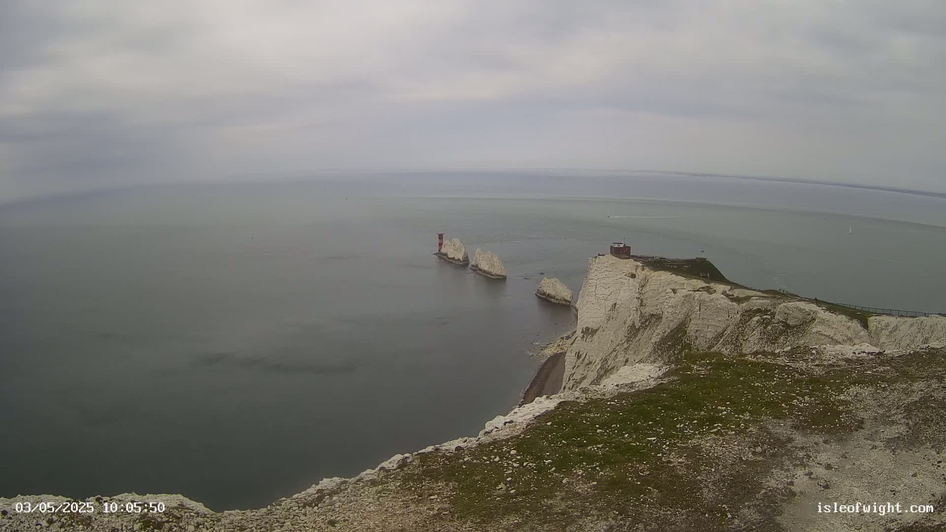 White cliffs overlook a calm, gray sea with three stacks of rock and a small building visible in the distance under an overcast sky.