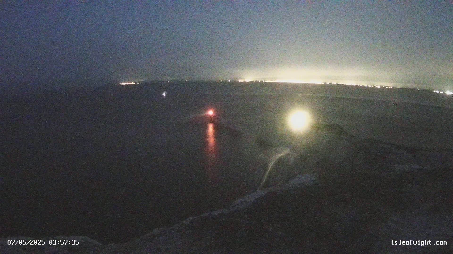 A dark, nighttime coastal scene shows a body of water with faint lights on the horizon and two small, illuminated objects in the water, one reddish and one yellowish.