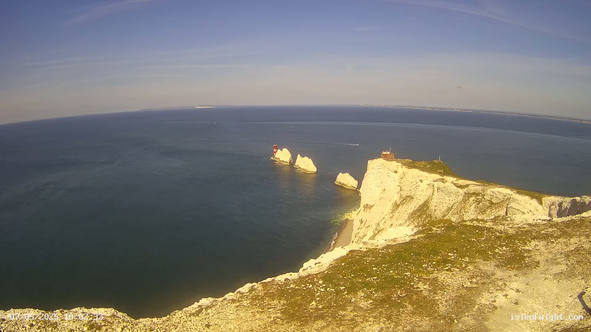A high-angle view shows white cliffs meeting a dark blue sea under a clear, sunny sky; three white rock formations jut out from the water near the cliffs, with a small building on one of the cliffs.