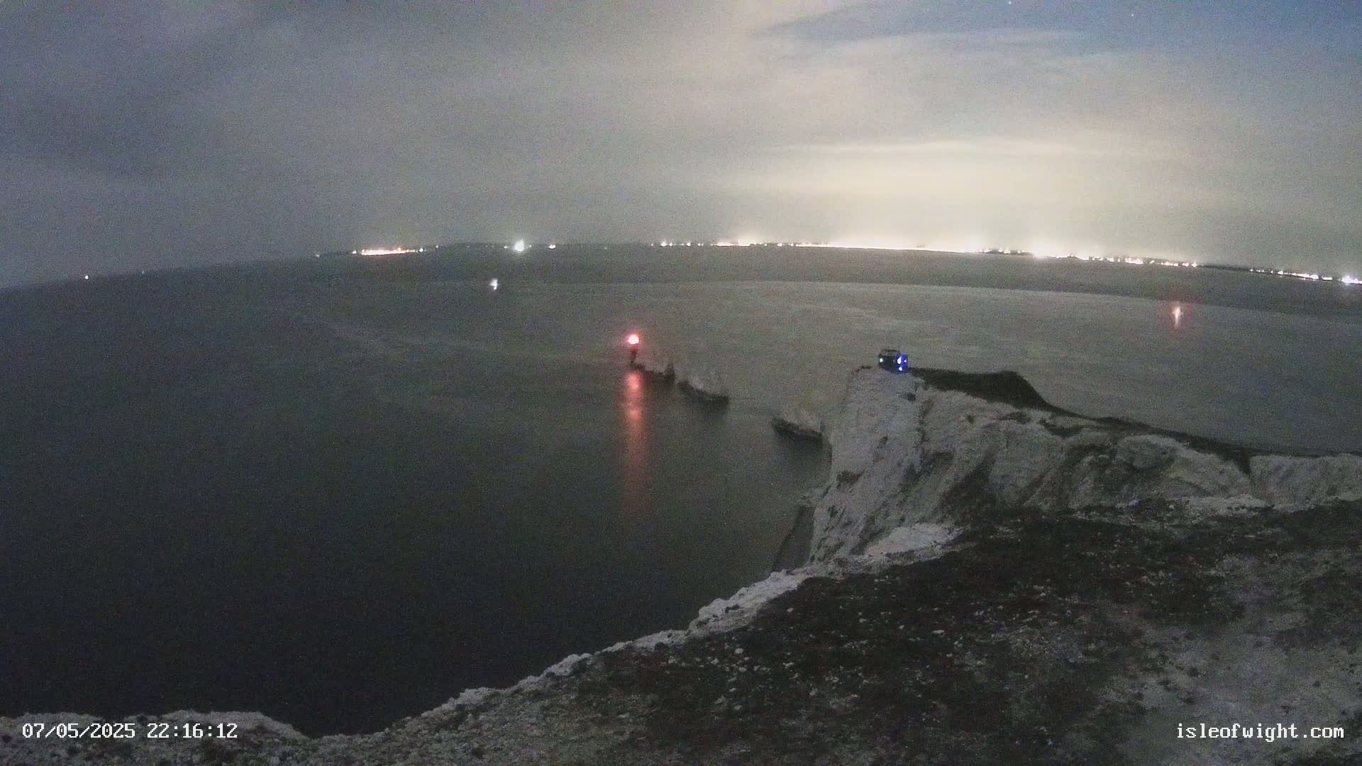 A nighttime view from a clifftop overlooking a dark sea with a lit lighthouse and distant city lights under a cloudy sky.