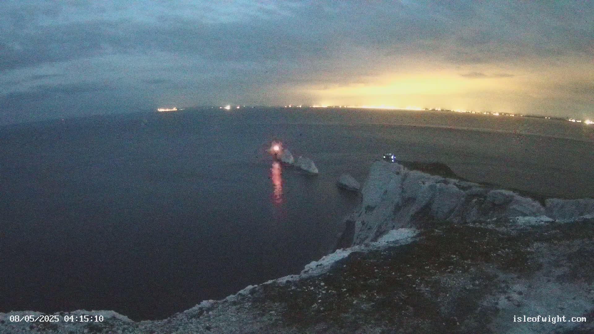 A nighttime view from a clifftop overlooking a dark sea with a distant city glowing on the horizon, and a lit structure on a rock formation in the water.