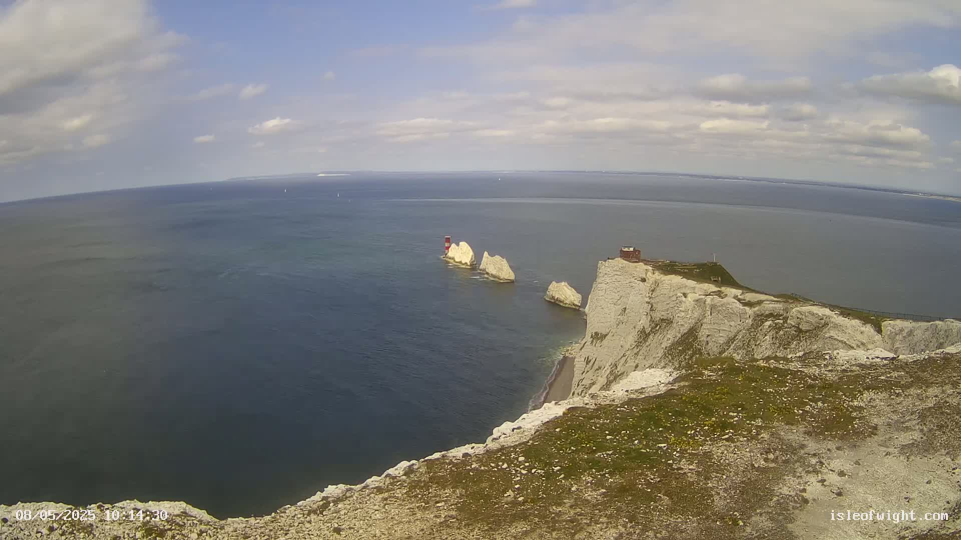 A high-angle view shows a partly cloudy sky over a calm sea with white cliffs and several stacks, one topped with a small red and white structure.