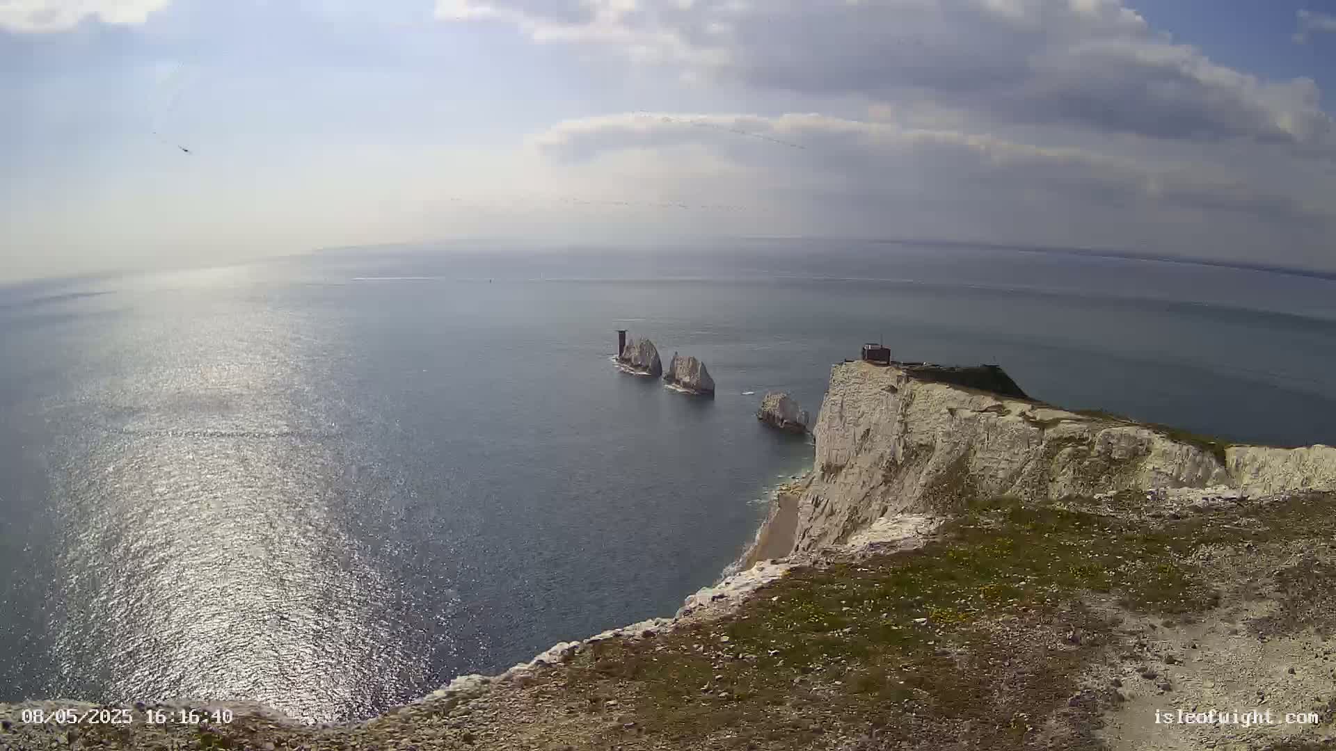 A high-angle view shows white cliffs overlooking a calm, sunlit sea with several small white islands in the distance under a partly cloudy sky.