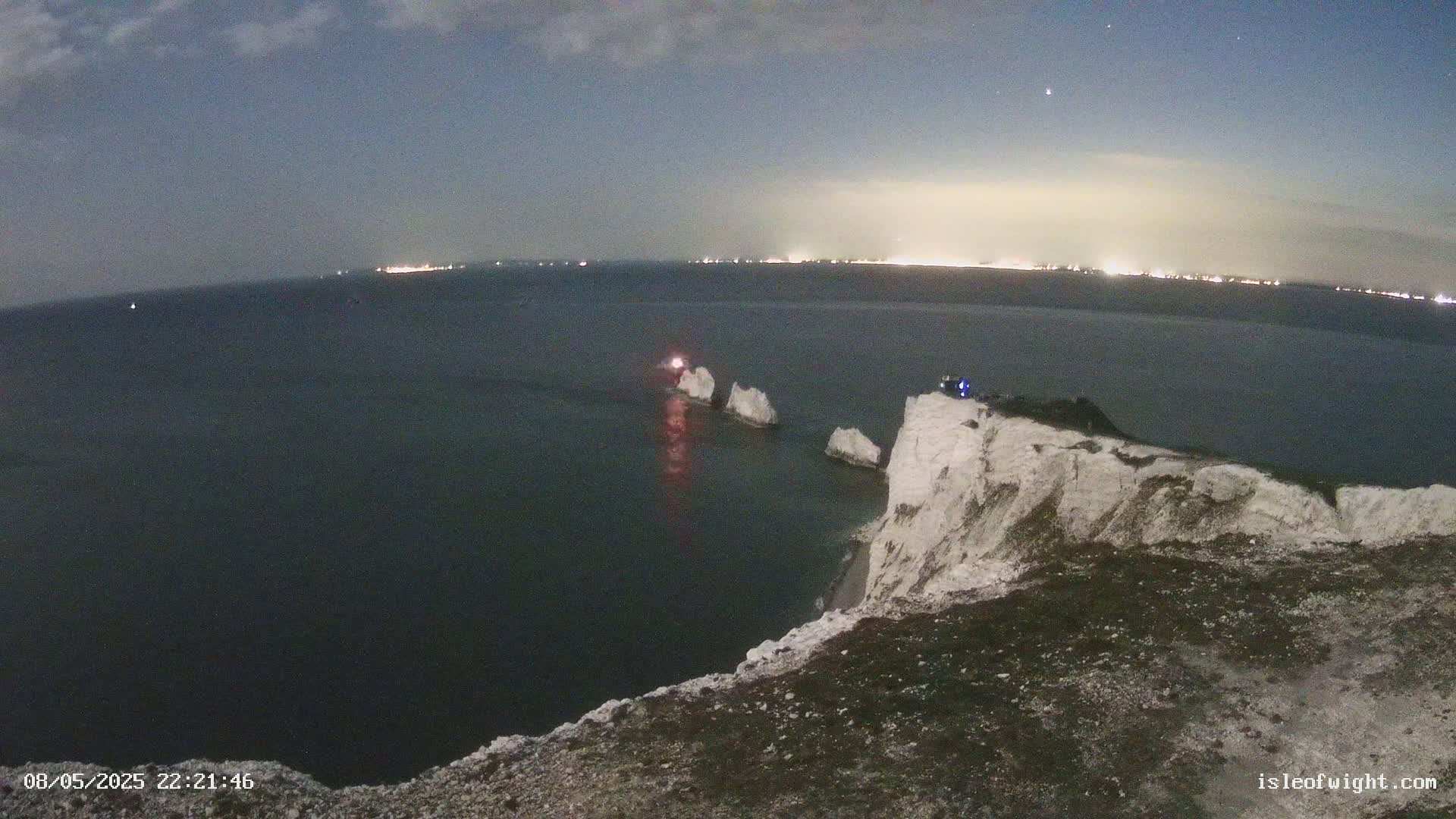 A nighttime view from a clifftop overlooking a dark sea with three white rock formations and a distant city skyline illuminated under a mostly clear sky.