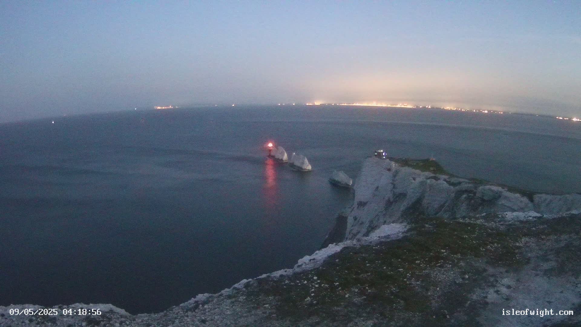 A high vantage point overlooks a calm, dark sea at twilight, showing two white rock formations with a lit beacon in the water between them, and a distant city skyline faintly illuminated on the horizon.