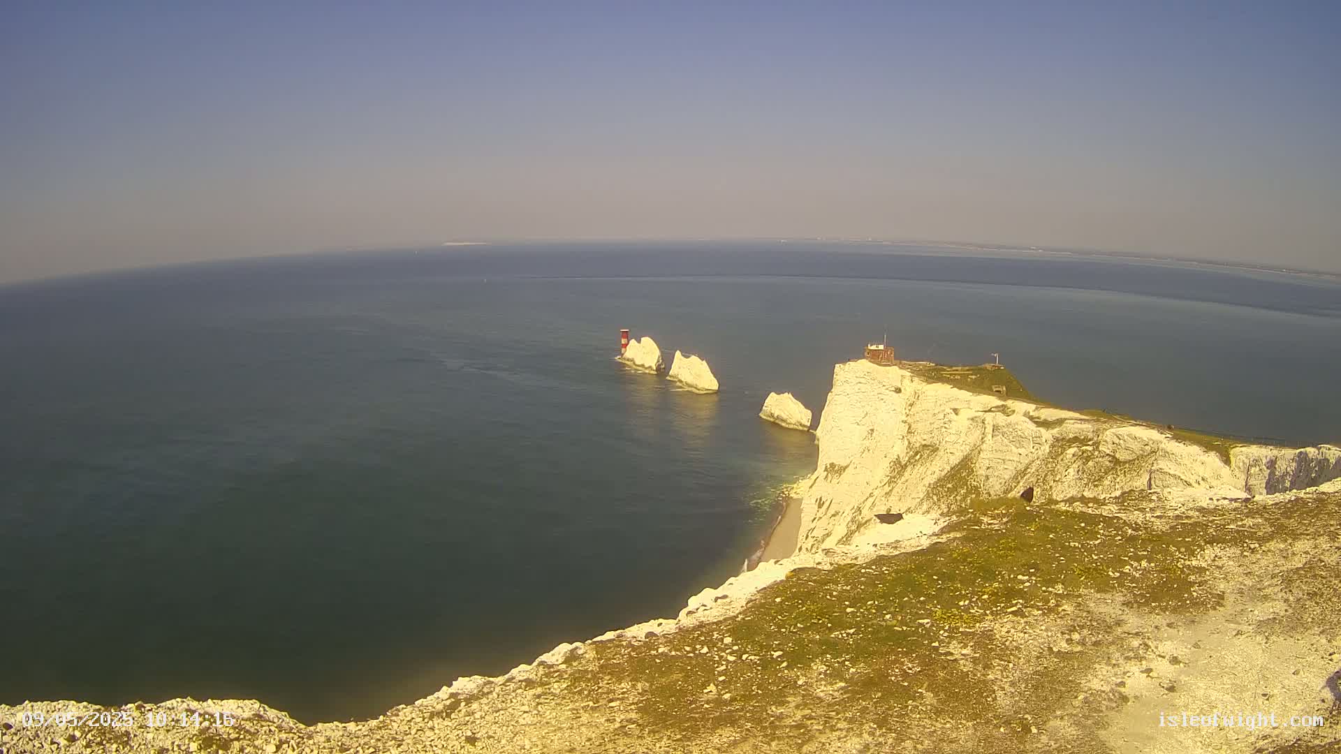 A high-angle view shows a cliff overlooking a calm sea with several small white rock formations and a small red building on the edge of the cliff on a clear, sunny day.